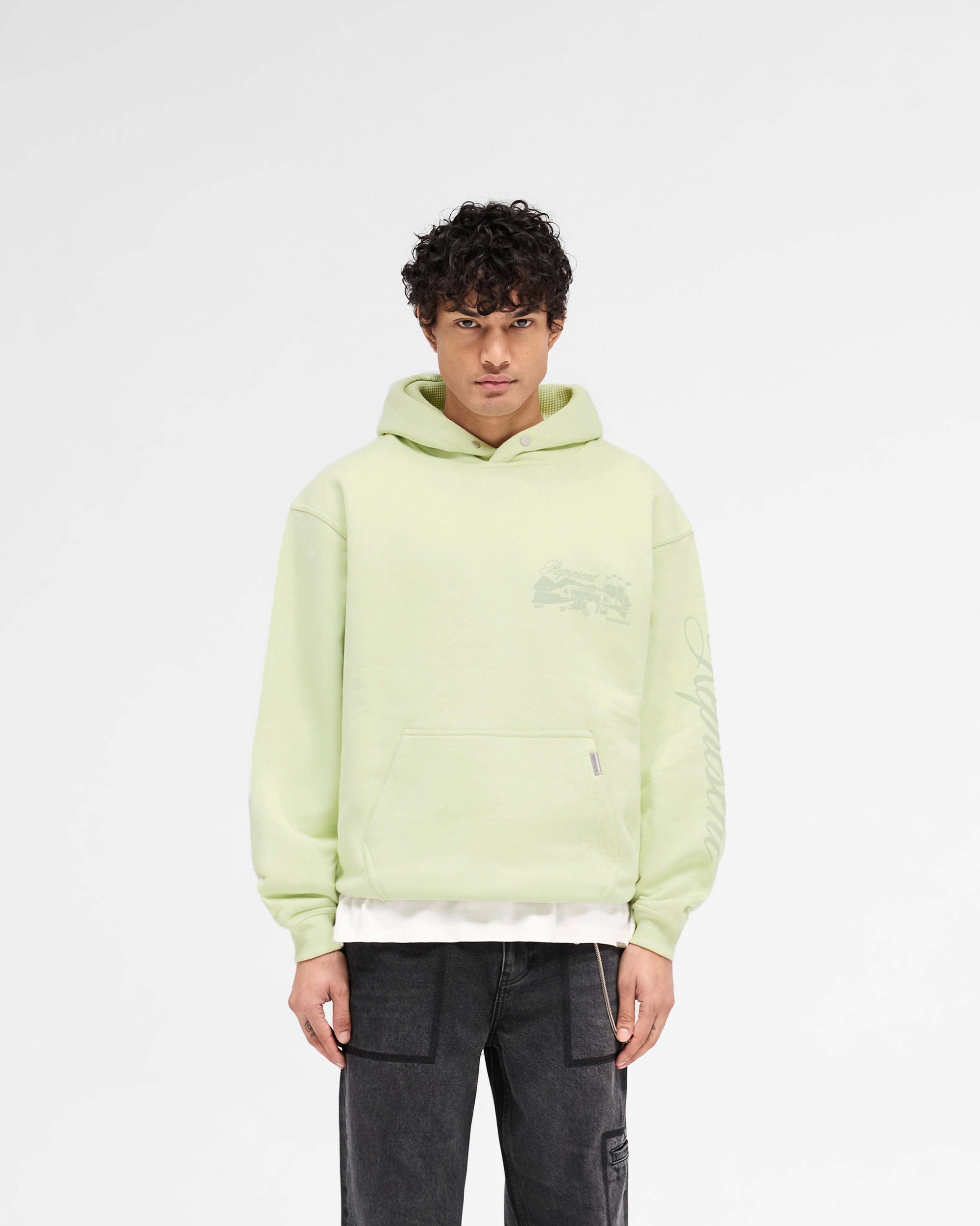 Man wearing a slightly oversized green hoodie on a plain white background | Raceway Hoodie in Matcha | Heavy weight jersey cotton hoodie with puff print graphics | SS25 Hoodies | Represent Clo