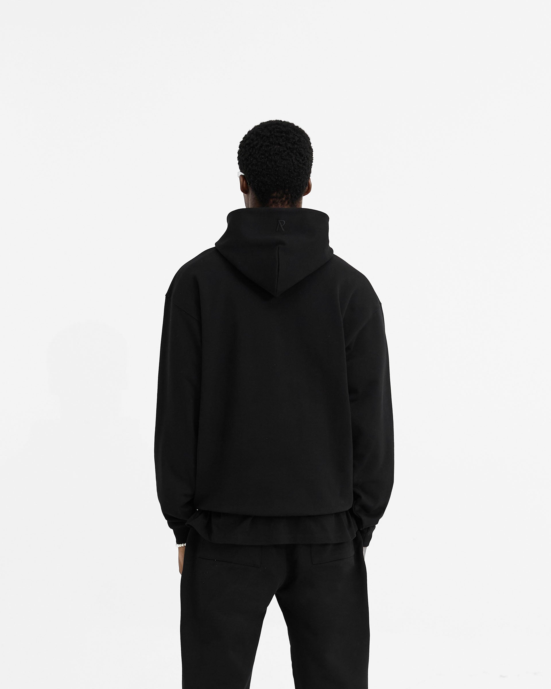 Man wearing a black oversized hoodie on a plain white background | Initial Oversized Hoodie in Black | Heavyweight hoodie with kangaroo pocket and embroidered details | Initial Hoodies | Represent Clo
