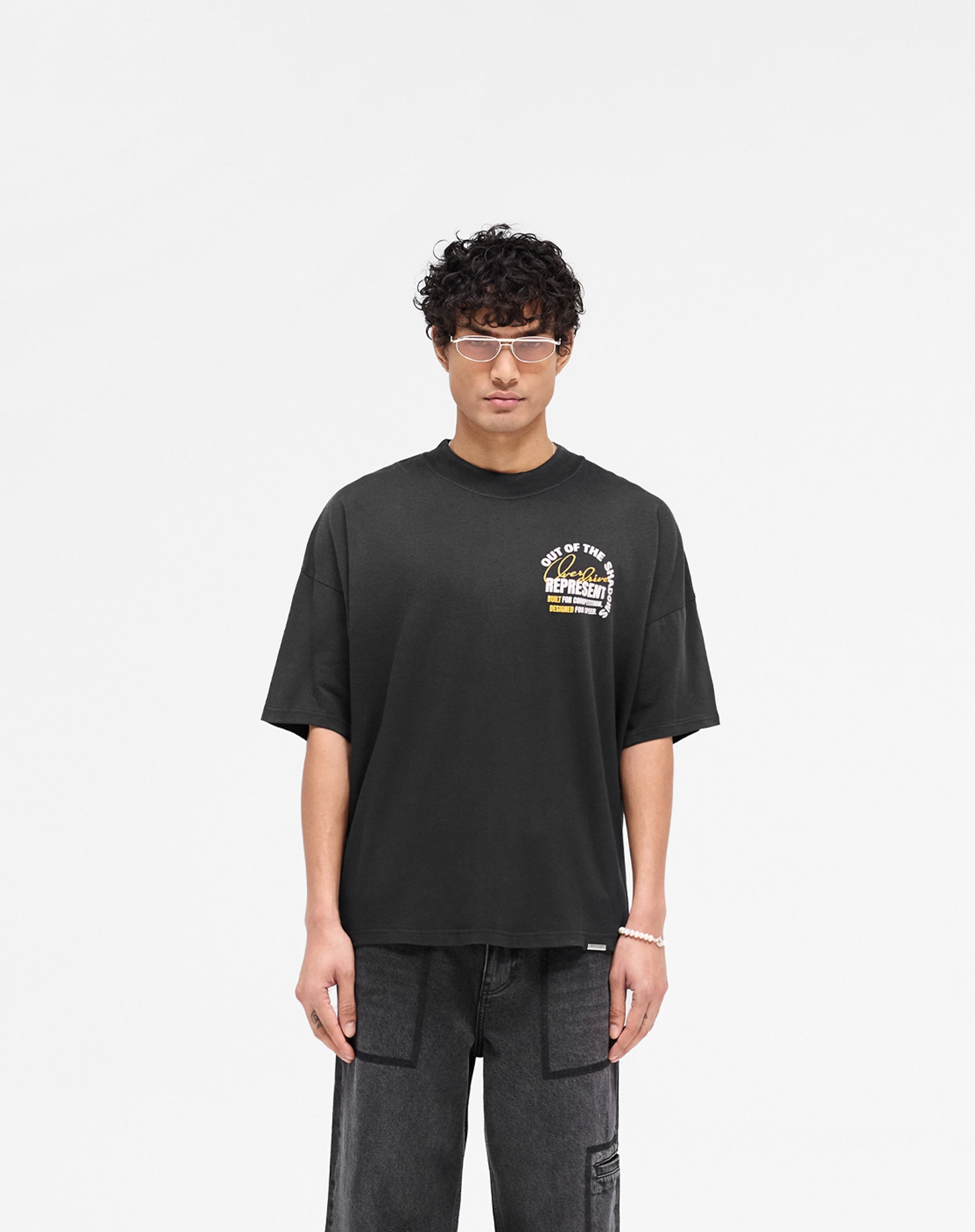 Man wearing a black graphic t-shirt on a plain white background | Out Of The Shadows T-Shirt in Stained Black | Boxy fit with fade out wash and metal bar hem | SS25 T-Shirts | Represent Clo