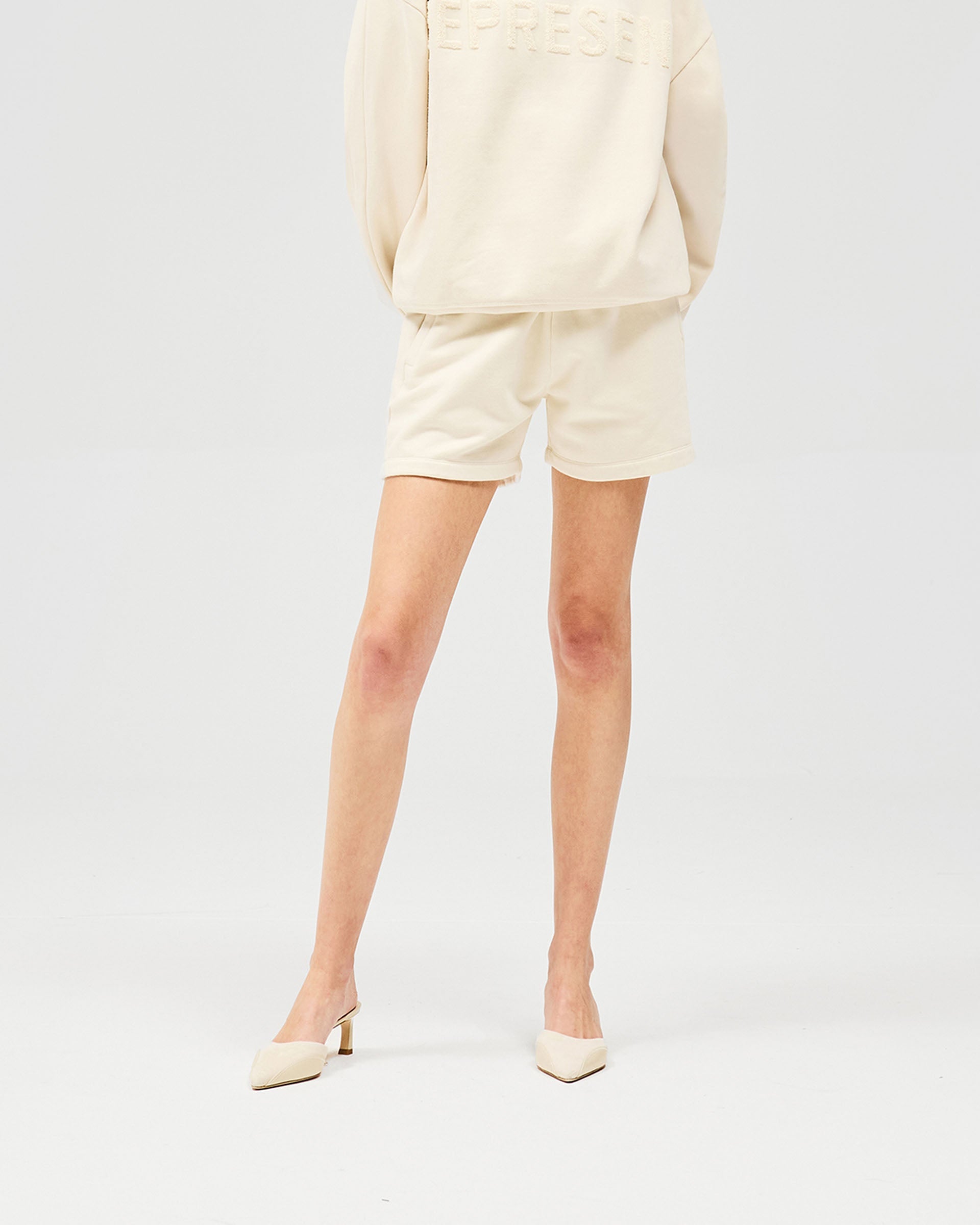 Woman wearing oat coloured mini shorts on a plain white background | Owners Club Short in Oat Milk | Sweat shorts with internal drawcord and side pockets | Womens Owners Club Shorts | Represent Clo