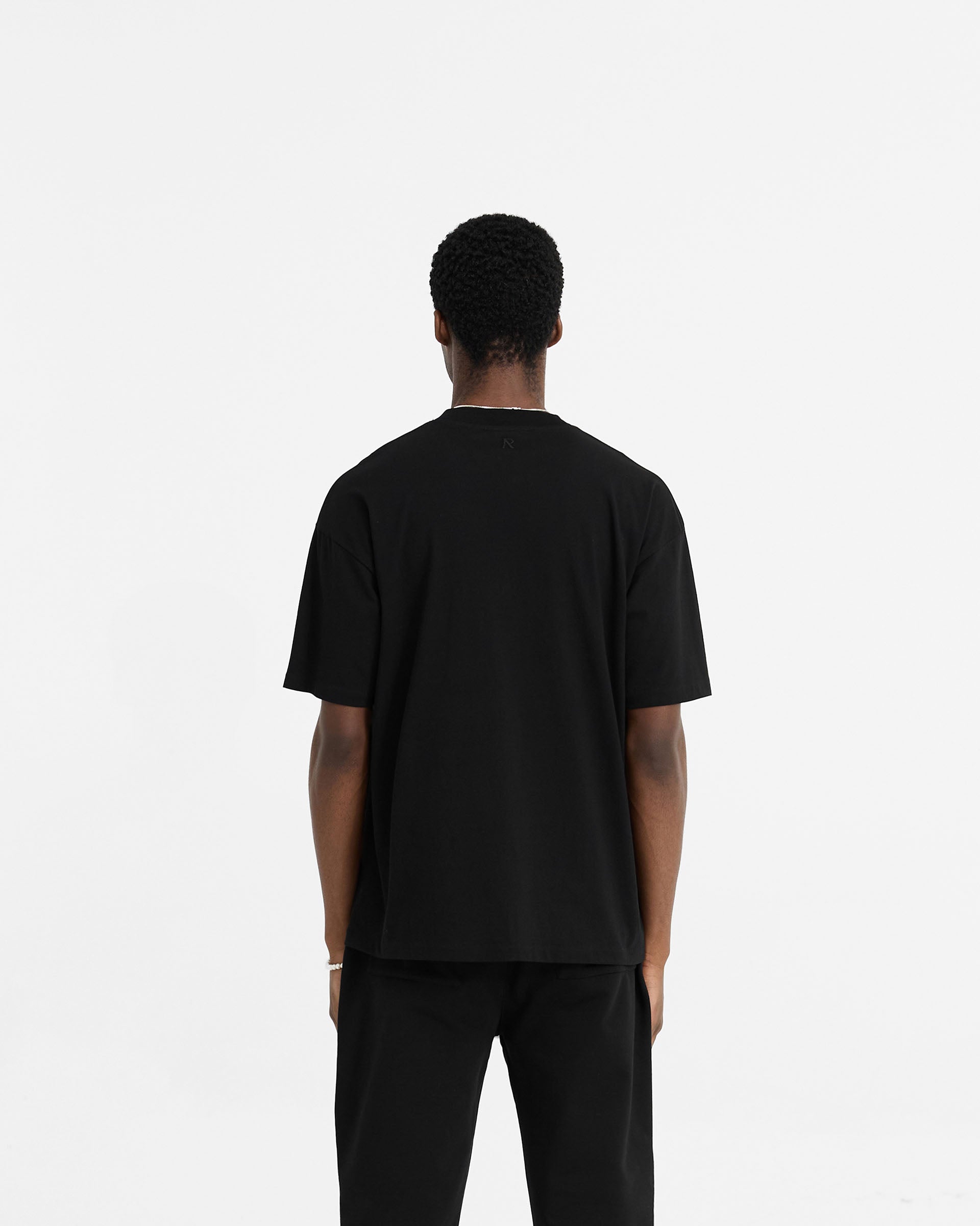 Man wearing a black oversized t-shirt on a plain white background | Initial Oversized T-Shirt in Black | Oversized t-shirt with embroidered details | Initial T-Shirts | Represent Clo
