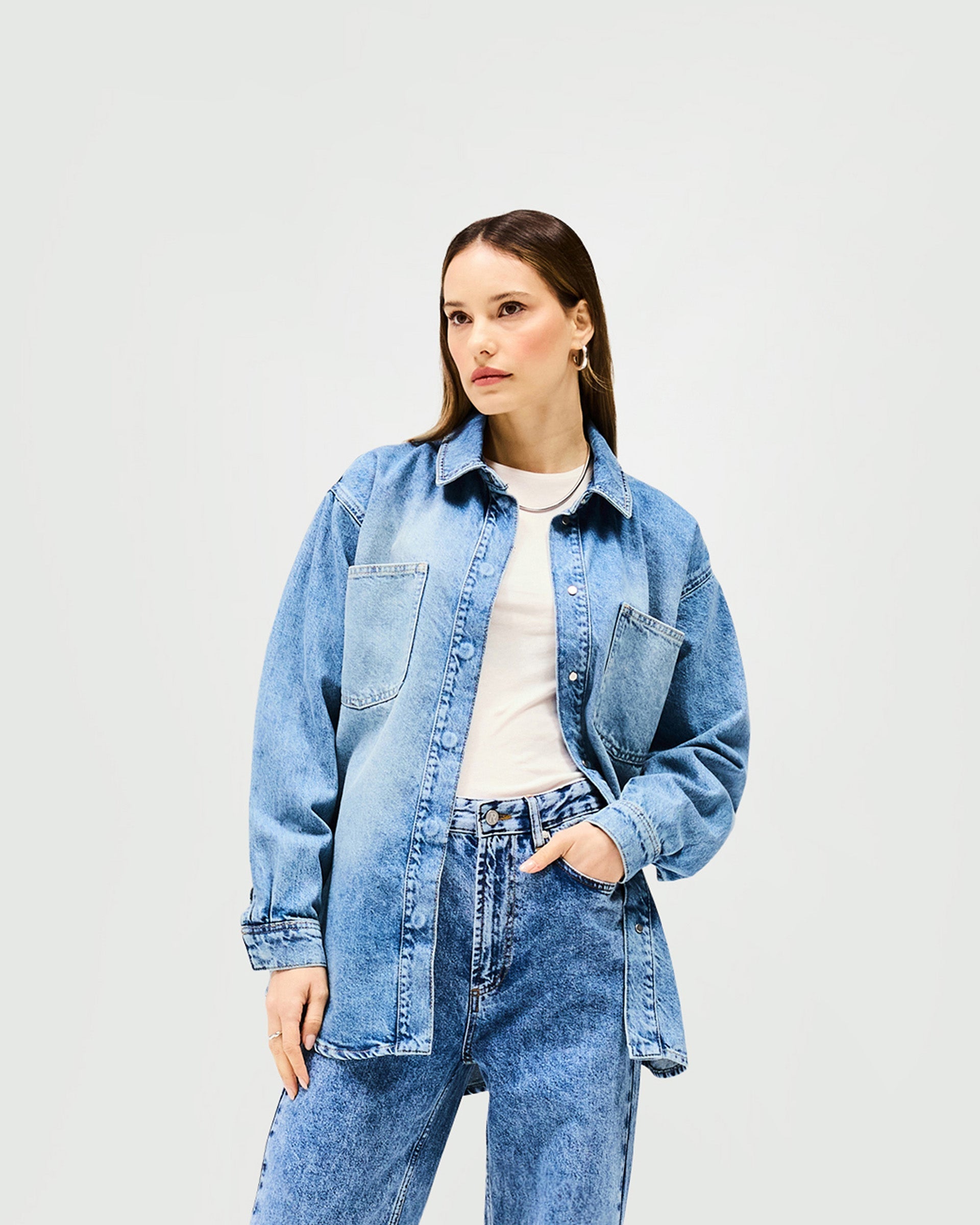 Woman wearing an oversized blue denim overshirt on a plain white background | Denim Overshirt in Washed Blue | Denim overshirt with branded popper fastenings | Womens SS25 Denim | Represent Clo
