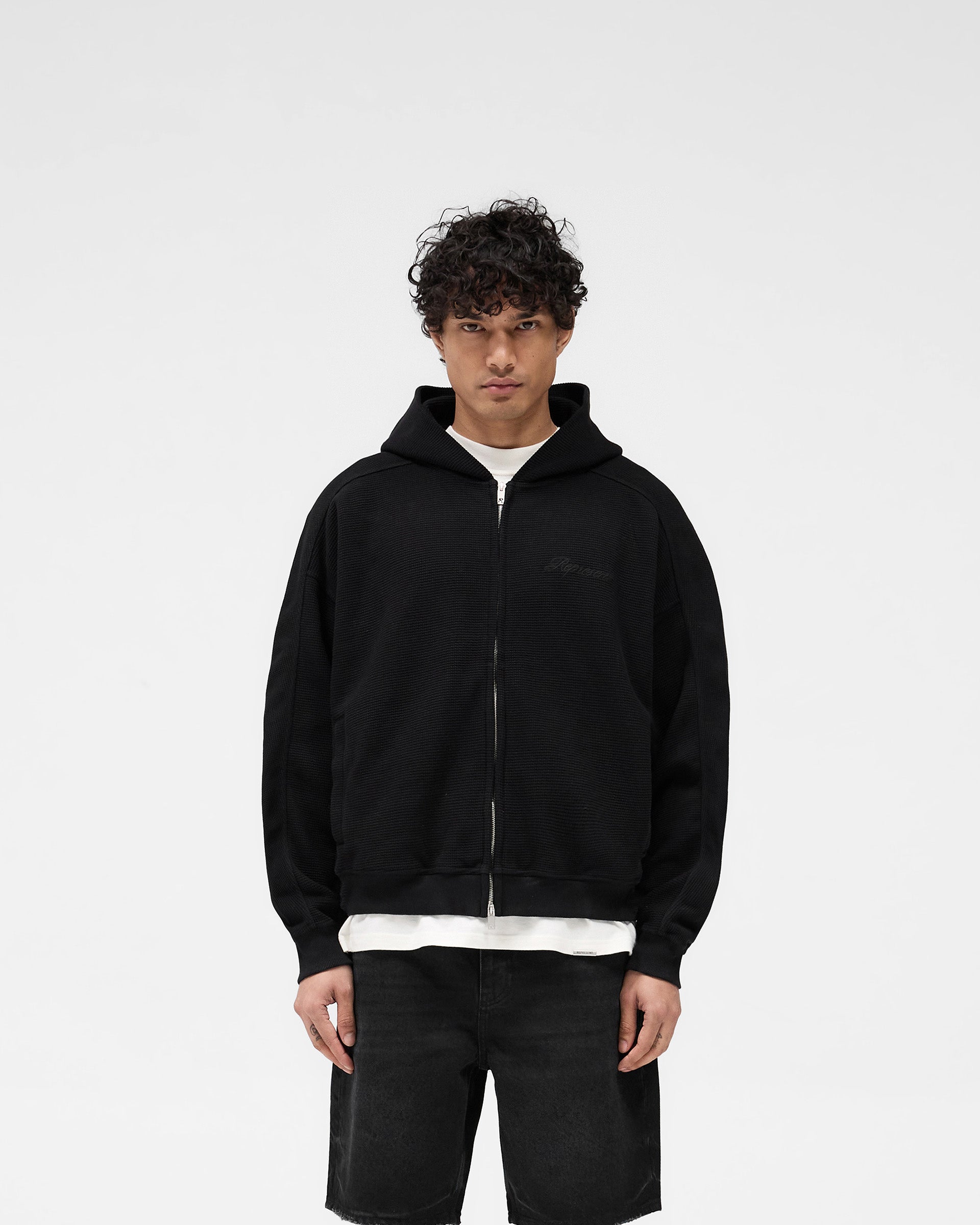 Man wearing a black oversized hoodie on a plain white background | Waffle Zip Through Hoodie in Jet Black | Boxy fit hoodie with chest embroidered logo and branded zipper | SS25 Hoodies | Represent Clo