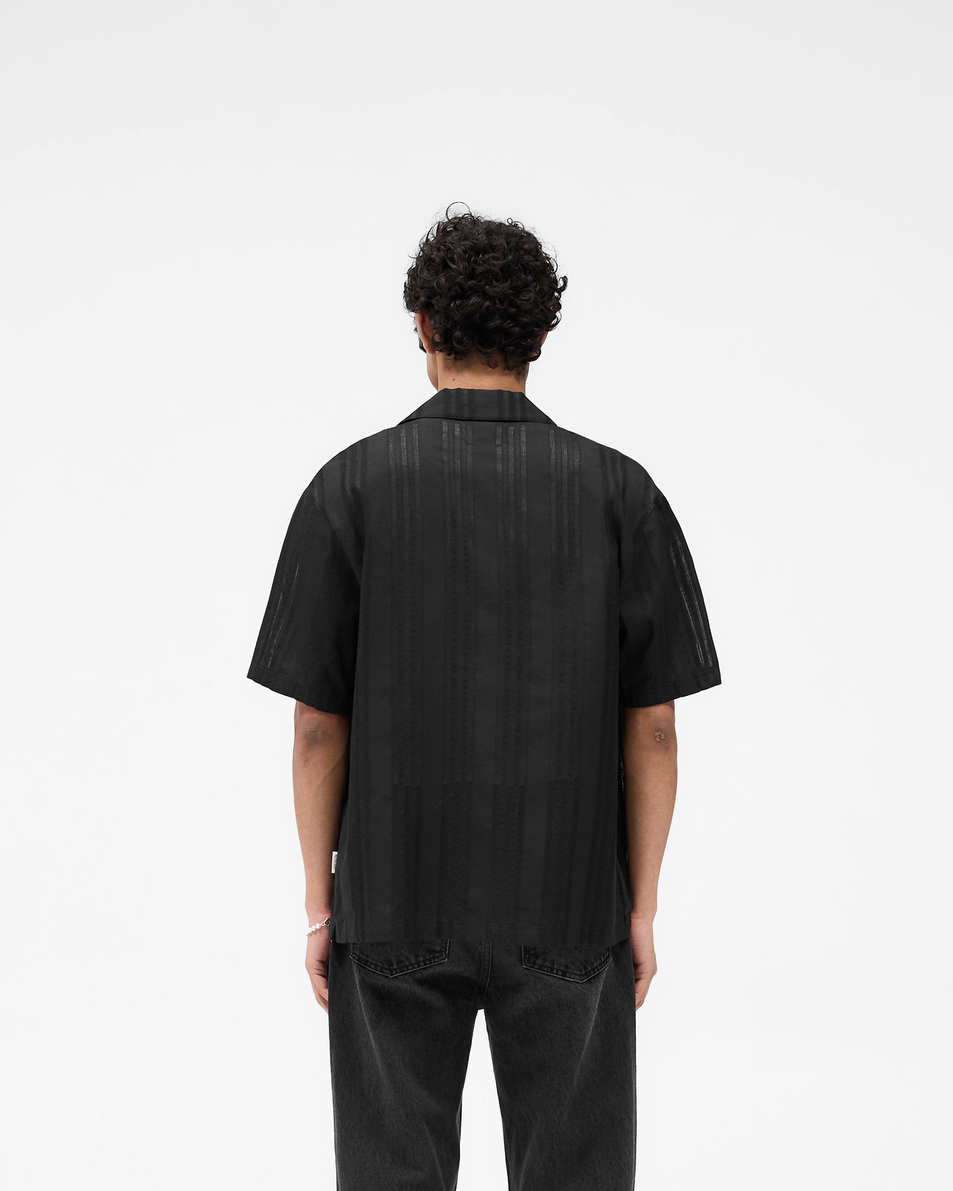 Man wearing a black embroidered bowling shirt on a plain white background | Embroidered Bowling Shirt in Jet Black | Sheer fabric with open weave and button-down front | SS25 Shirts | Represent Clo