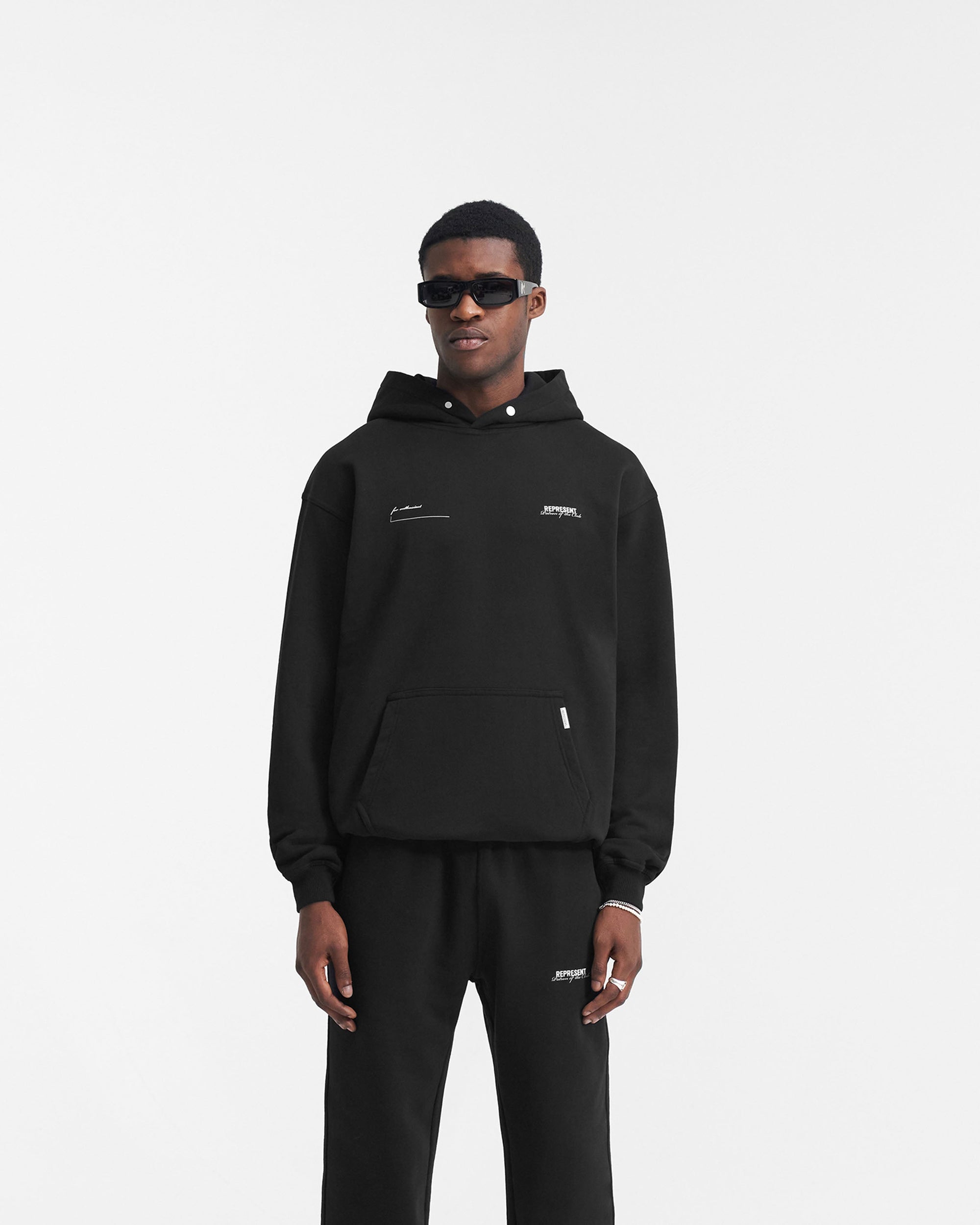 Man wearing a black oversized hoodie on a plain white background | Patron Of The Club Hoodie in Black | Heavyweight jersey cotton oversized hoodie | Patrons Hoodies | Represent Clo