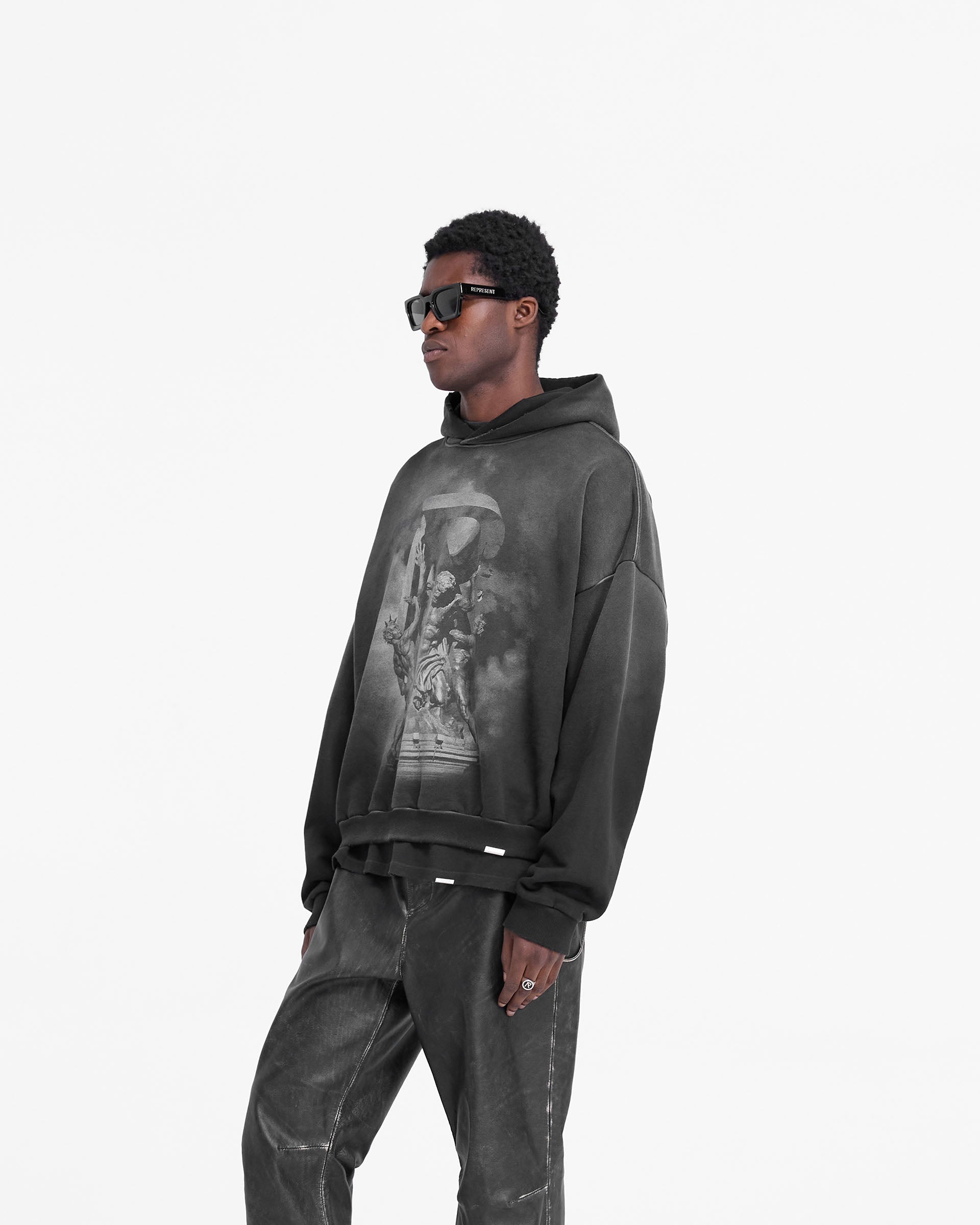 Man wearing an oversized black hoodie on a plain white background | Atlas Hoodie in Stained Black | Heavy weight jersey cotton with puff and screen print graphics | FW24 Hoodies | Represent Clo