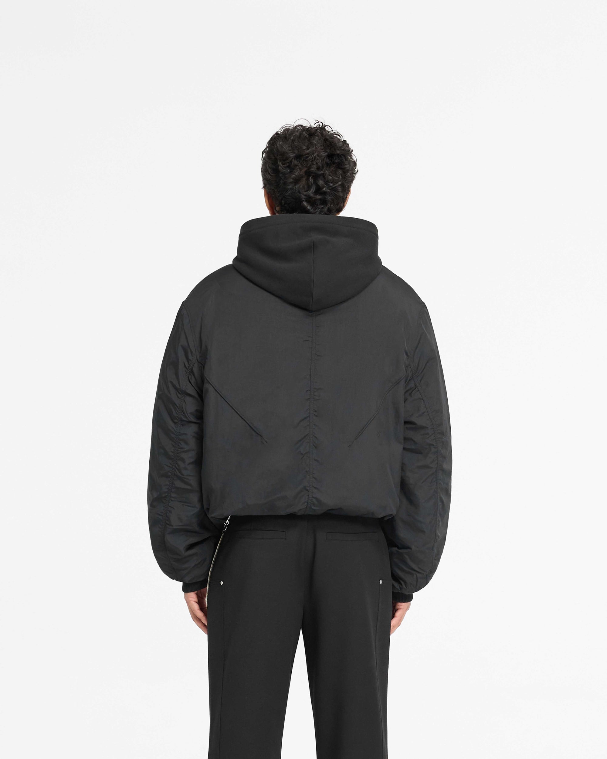 Man wearing a black bomber jacket with hood on a plain white background | Nylon Hooded Bomber Jacket in Black | Oversized nylon body with metal logo | PRE SS25 Outerwear | Represent Clo