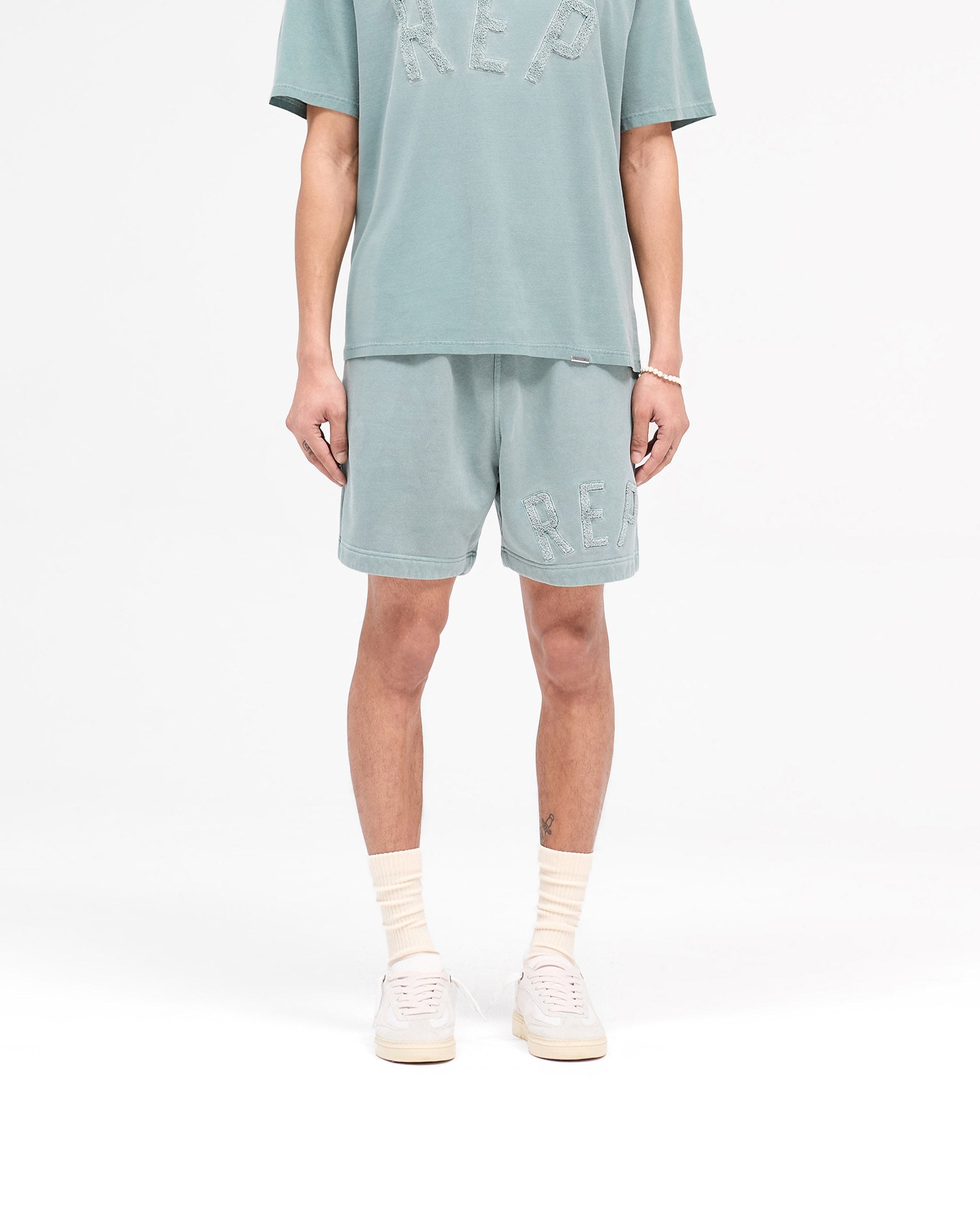 Man wearing green shorts on a plain white background | Rep Applique Shorts in Forest Green | Shorts with elasticated waistband and back pockets | SS25 Shorts | Represent Clo