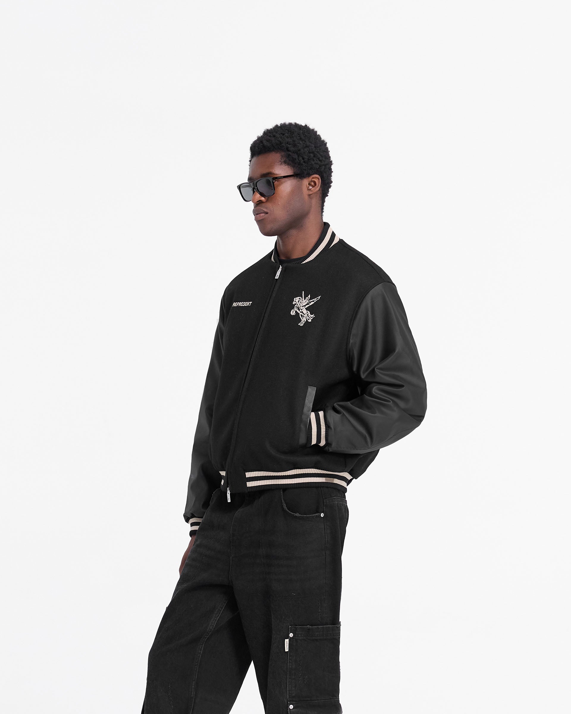Man wearing a black varsity jacket on a plain white background | Mascot Wool Varsity Jacket in Jet Black | Varsity jacket with embroidered mascot and PU sleeves | FW24 Outerwear | Represent Clo