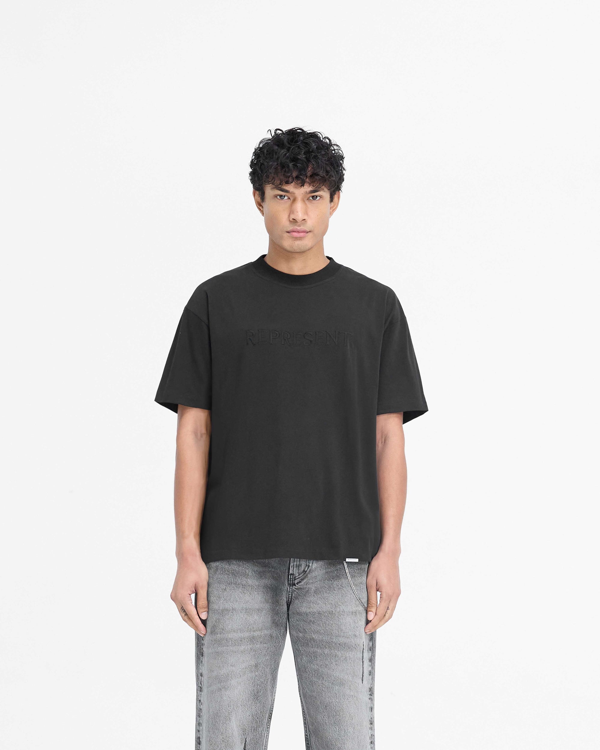 Man wearing a black t-shirt on a plain white background | Serif Logo T-Shirt in Jet Black | Slightly oversized t-shirt with high build embroidered serif branding | PRE SS25 T-Shirts | Represent Clo
