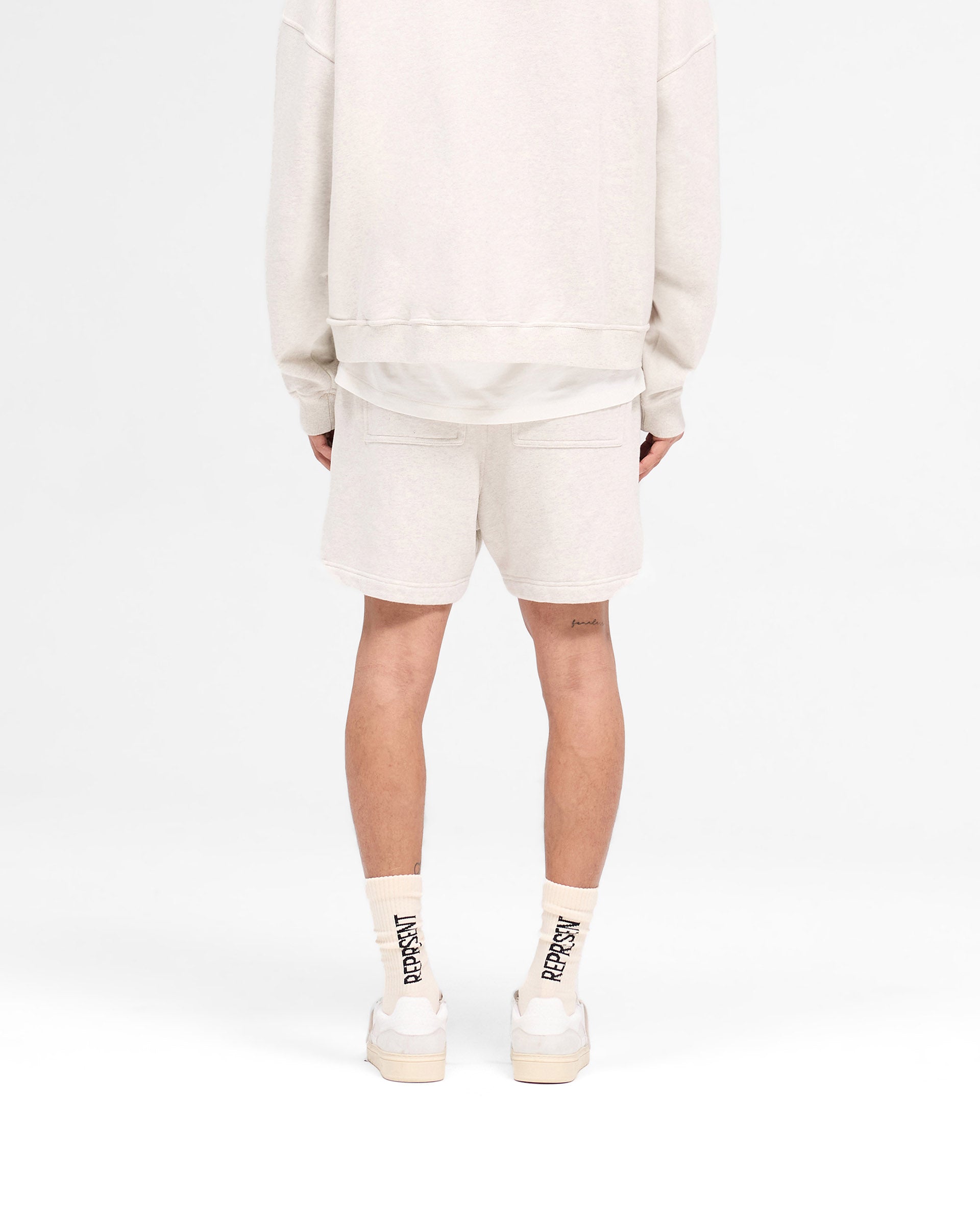 Man wearing cream relaxed shorts on a plain white background | Rep Applique Shorts in Cream Marl | Shorts with elasticated waistband and back pockets | SS25 Shorts | Represent Clo