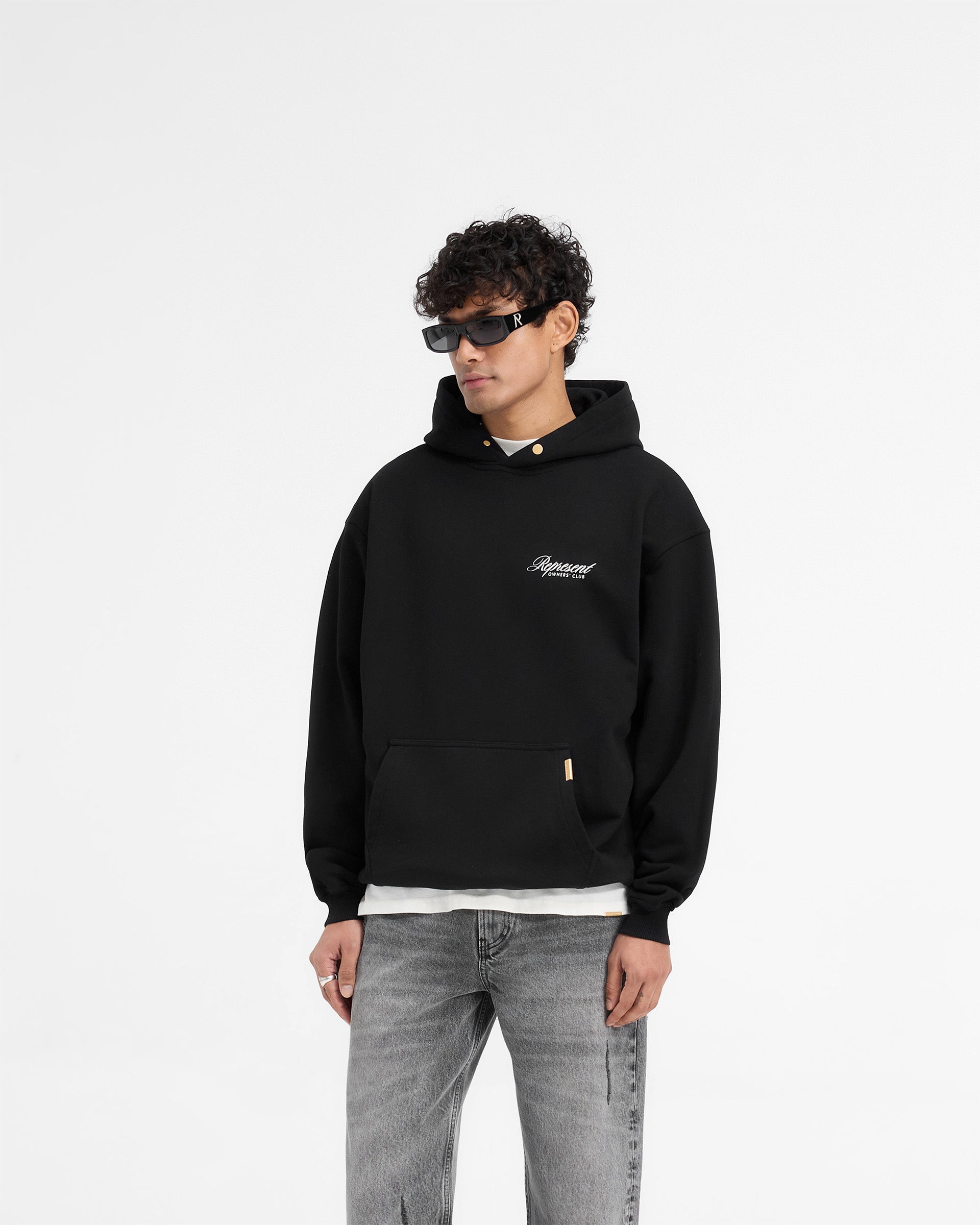 Man wearing a black oversized hoodie on a plain white background | Represent X Harrods Bear Owners Club Hoodie in Jet Black | Oversized hoodie with Harrods Bear graphic and gold detailing | Exclusives Hoodies | Represent Clo