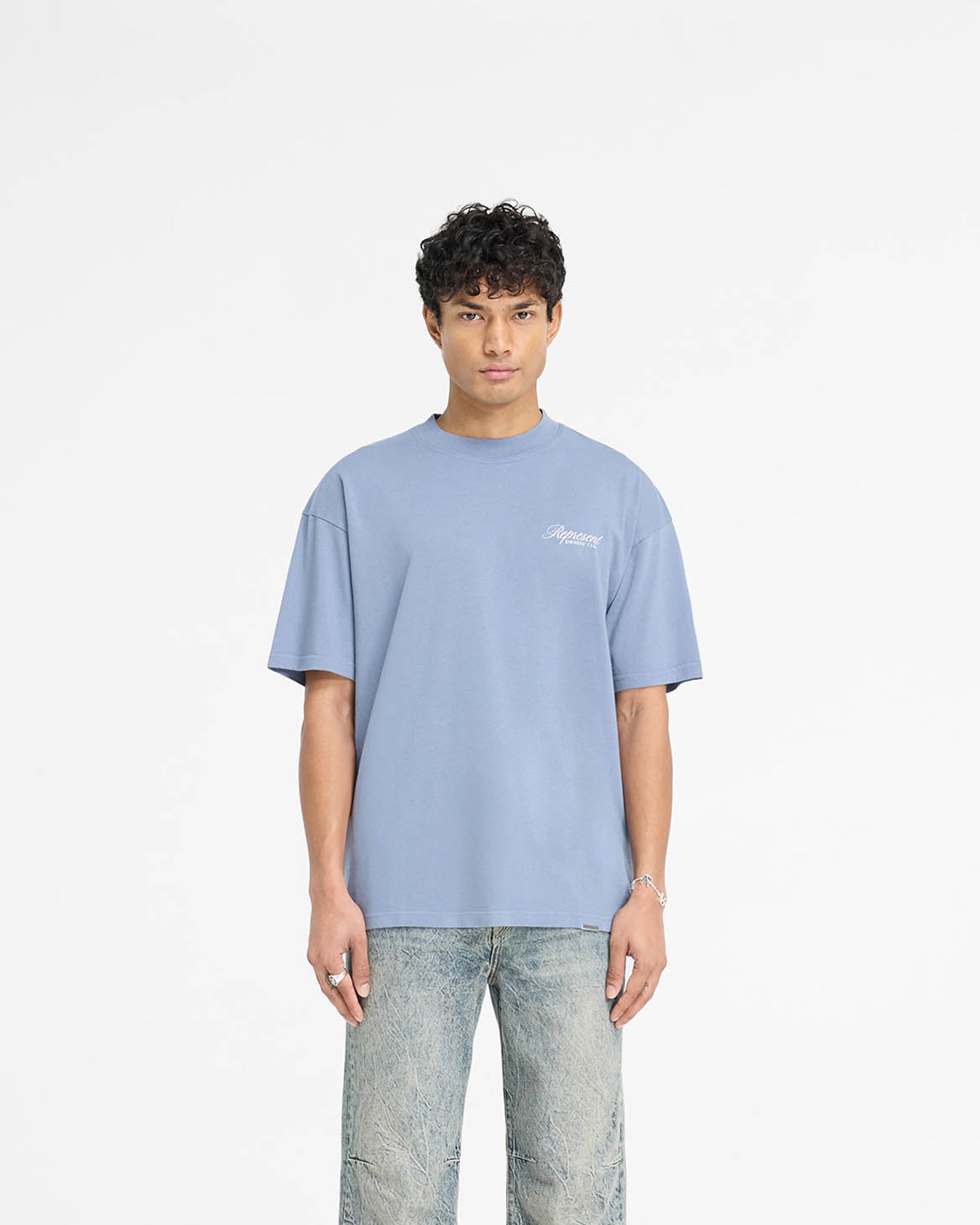 Man wearing an oversized blue t-shirt on a plain white background | Represent Owners Club Script T-Shirt in Dusty Blue | T-Shirt with ribbed crewneck and screen print | Owners Club T-Shirts | Represent Clo