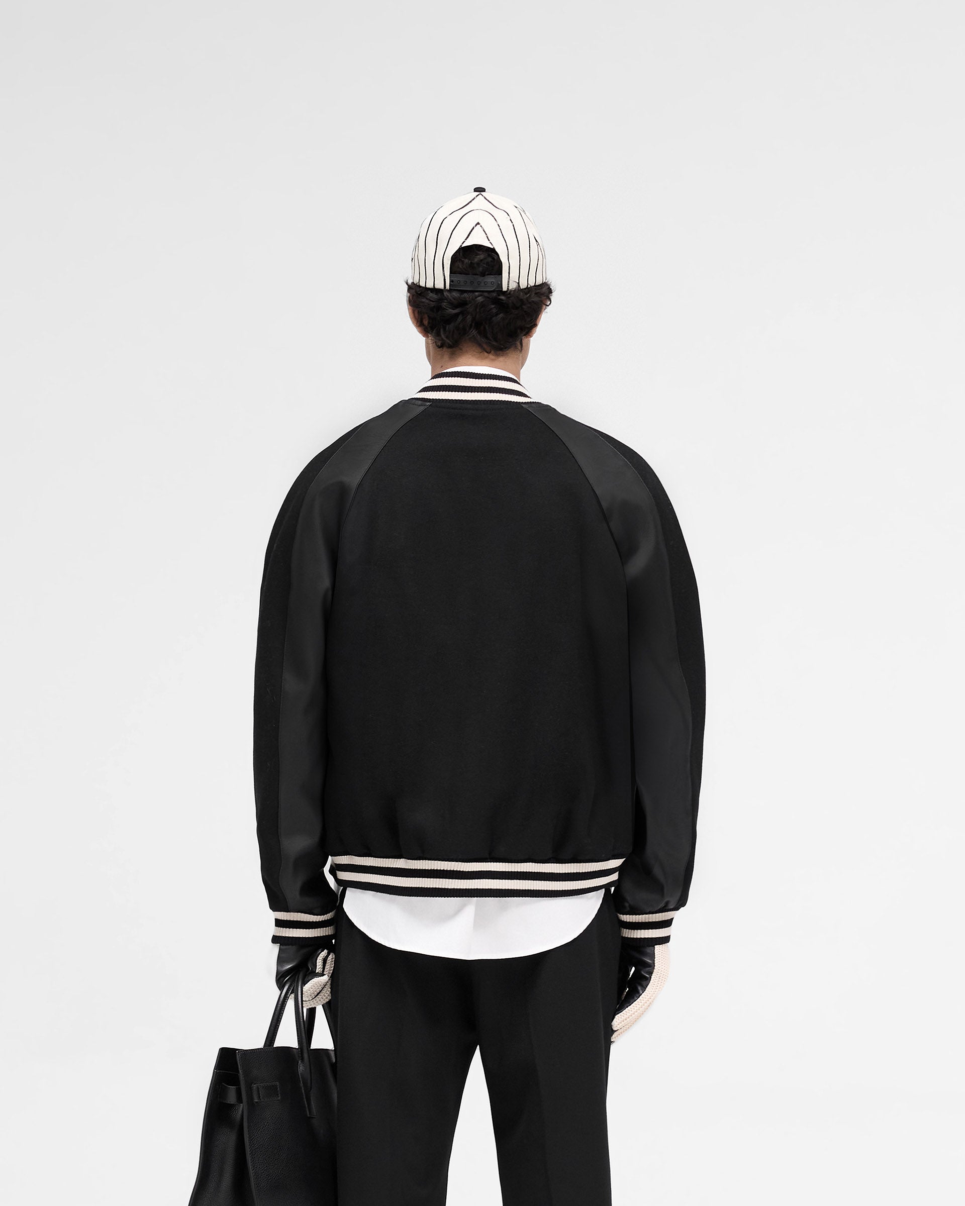 Man wearing a black raglan varsity jacket on a plain white background | Raglan Varsity Jacket in Black | Oversized fit with raglan sleeves and zip pockets | SS25 Outerwear | Represent Clo