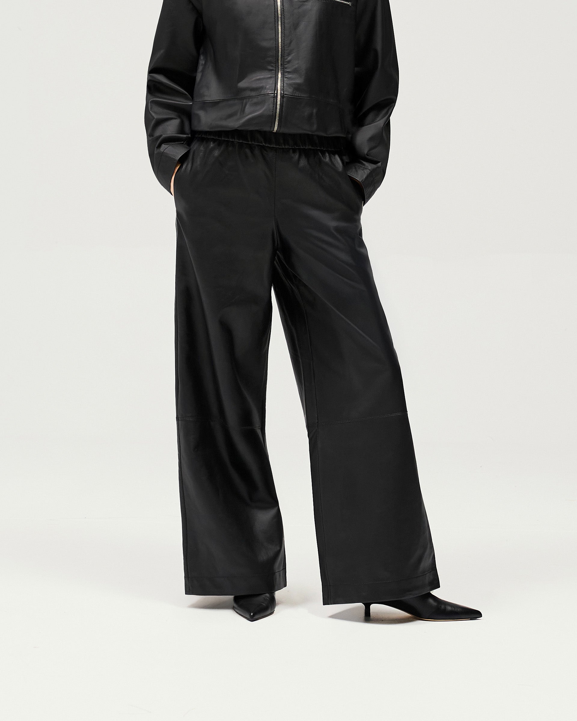 Woman wearing black leather track pants on a plain white background | Fuel Trouser in Jet Black | Relaxed fit wide leg trousers with elasticated waist | Womens SS25 Bottoms | Represent Clo