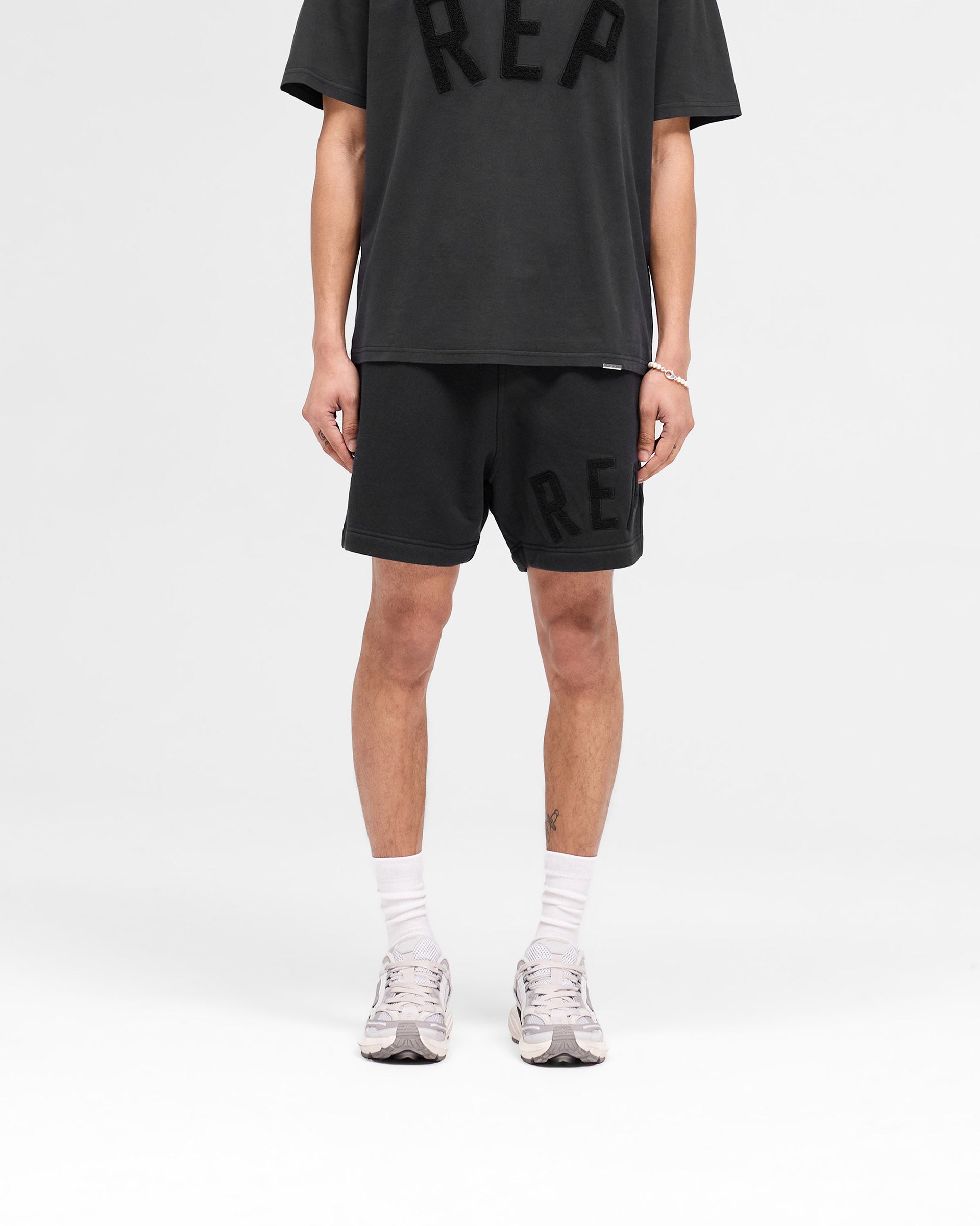 Man wearing off black shorts on a plain white background | Rep Applique Shorts in Off Black | Shorts with elasticated waistband and applique logo | SS25 Shorts | Represent Clo