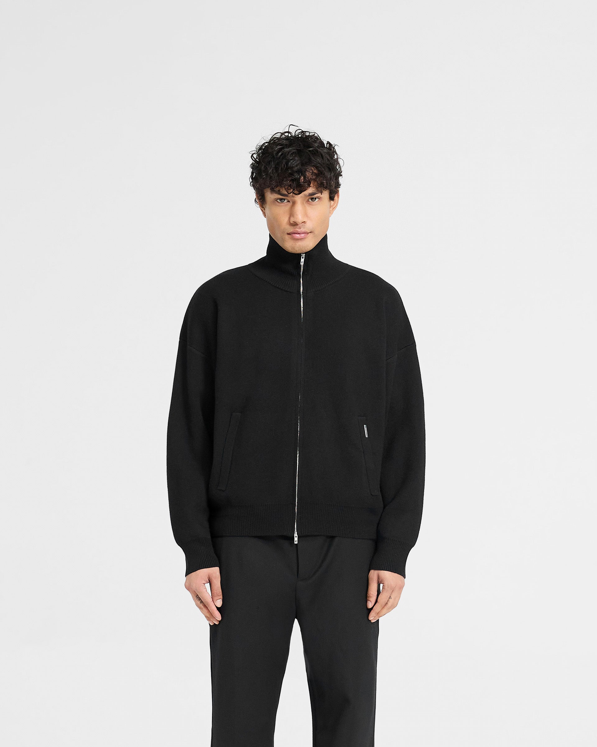 Man wearing a black knitted track jacket on a plain white background | Initial Knitted Track Jacket in Jet Black | Oversized track jacket with zip closure | Initial Knitwear | Represent Clo