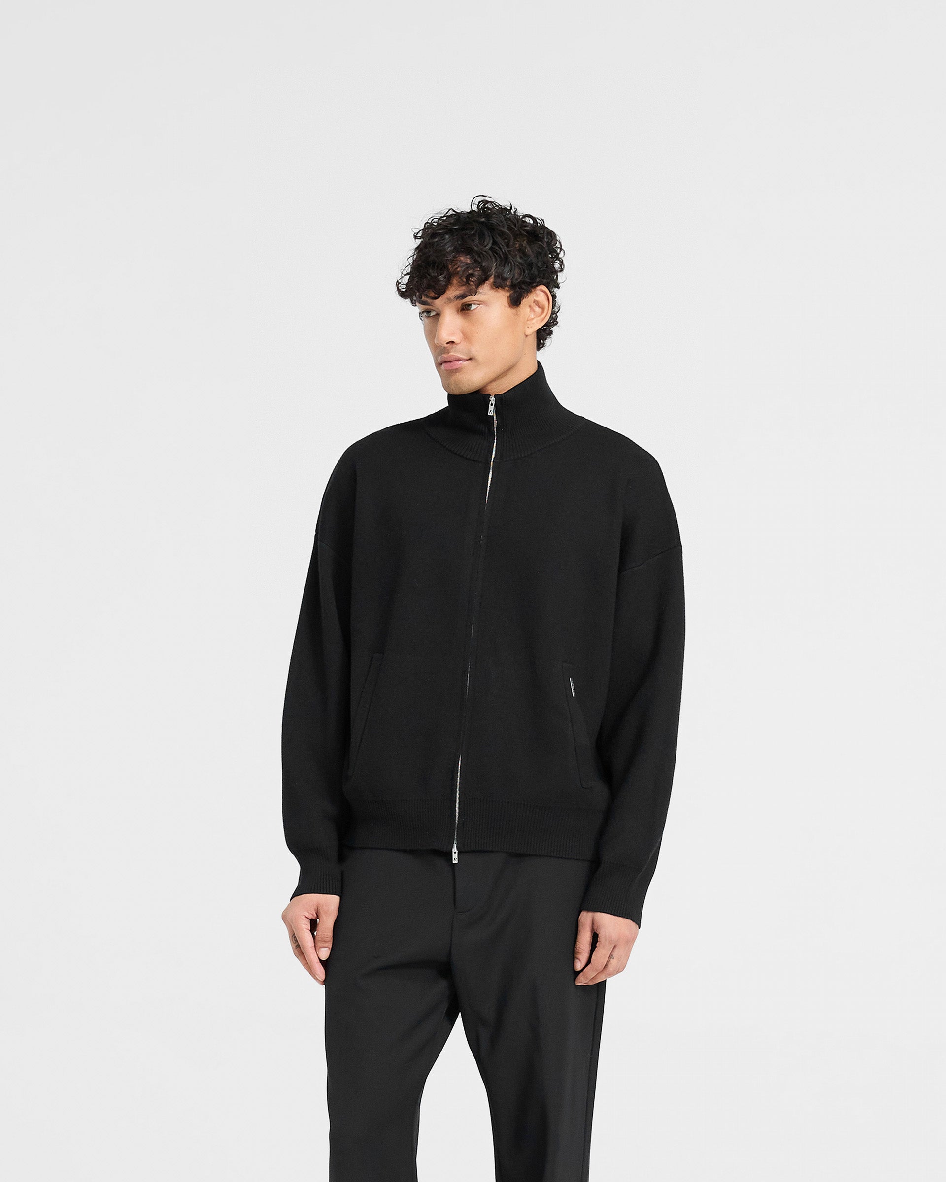 Man wearing a black knitted track jacket on a plain white background | Initial Knitted Track Jacket in Jet Black | Knitted track jacket with double ended zip and oversized fit | Initial Knitwear | Represent Clo