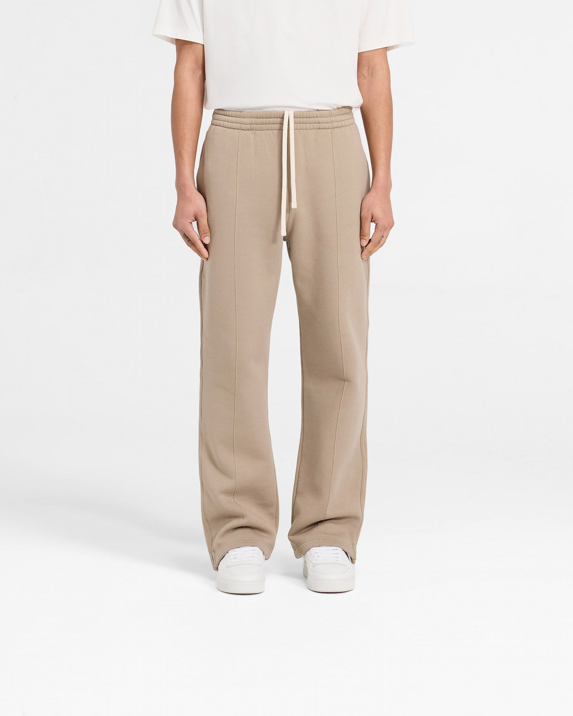 Man wearing beige wide leg sweatpants on a plain white background | Initial Sweatpants in Dune | Sweatpants with pintuck detail and split hems | Initial Pants | Represent Clo