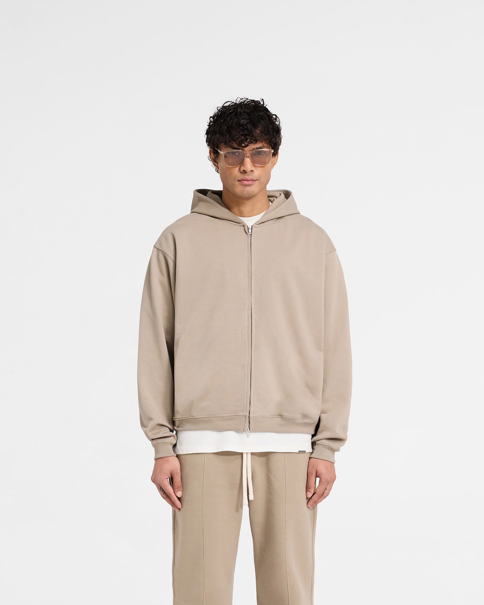 Man wearing an oversized beige hoodie on a plain white background | Initial Zip Hoodie in Dune | Loopback jersey cotton hoodie with zip closure | Initial Hoodies | Represent Clo