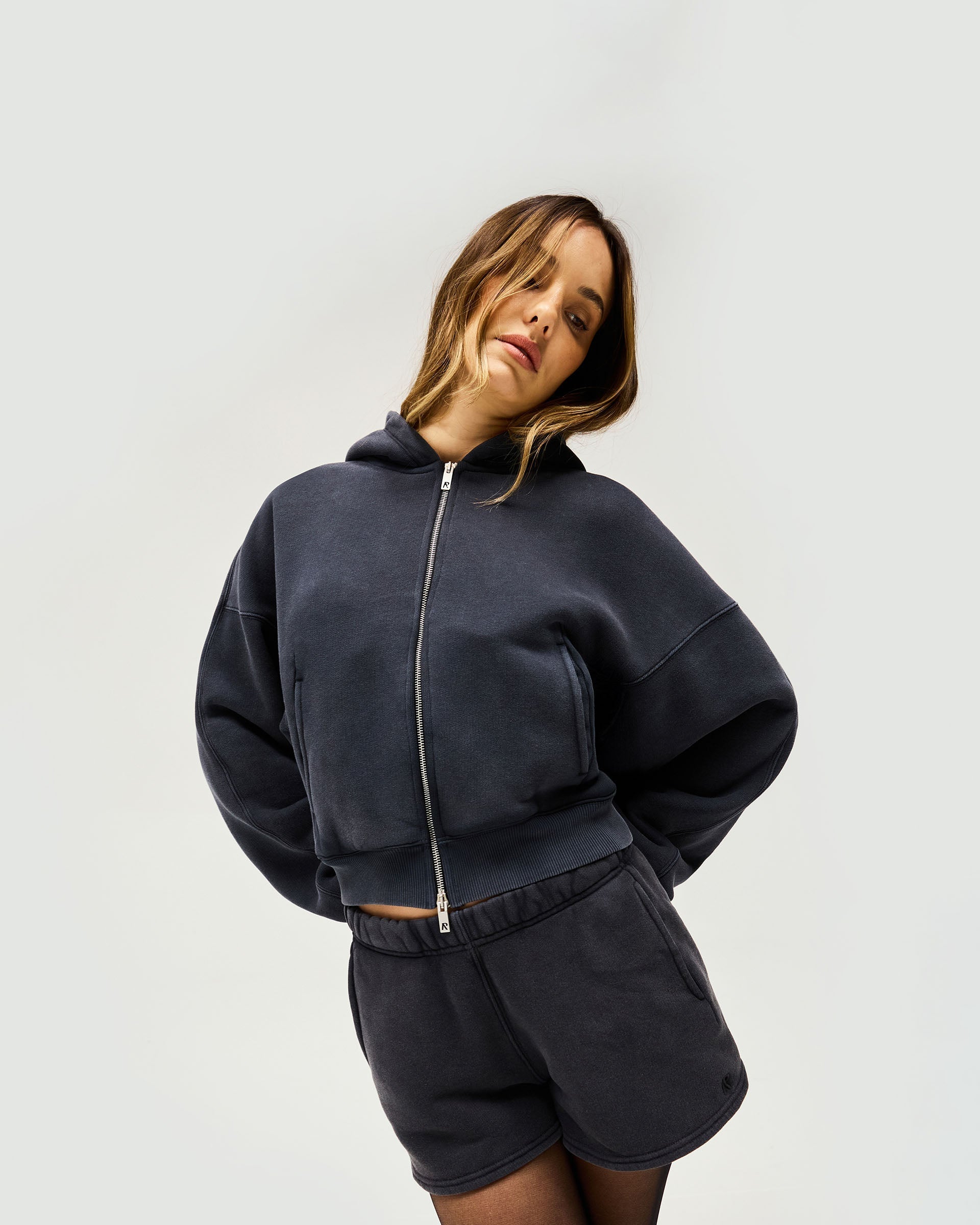 Woman wearing an oversized black hoodie on a plain white background | Initial Oversized Crop Hoodie in Washed Black | Cropped fit hoodie with branded metal zip | Womens Initial Hoodies | Represent Clo