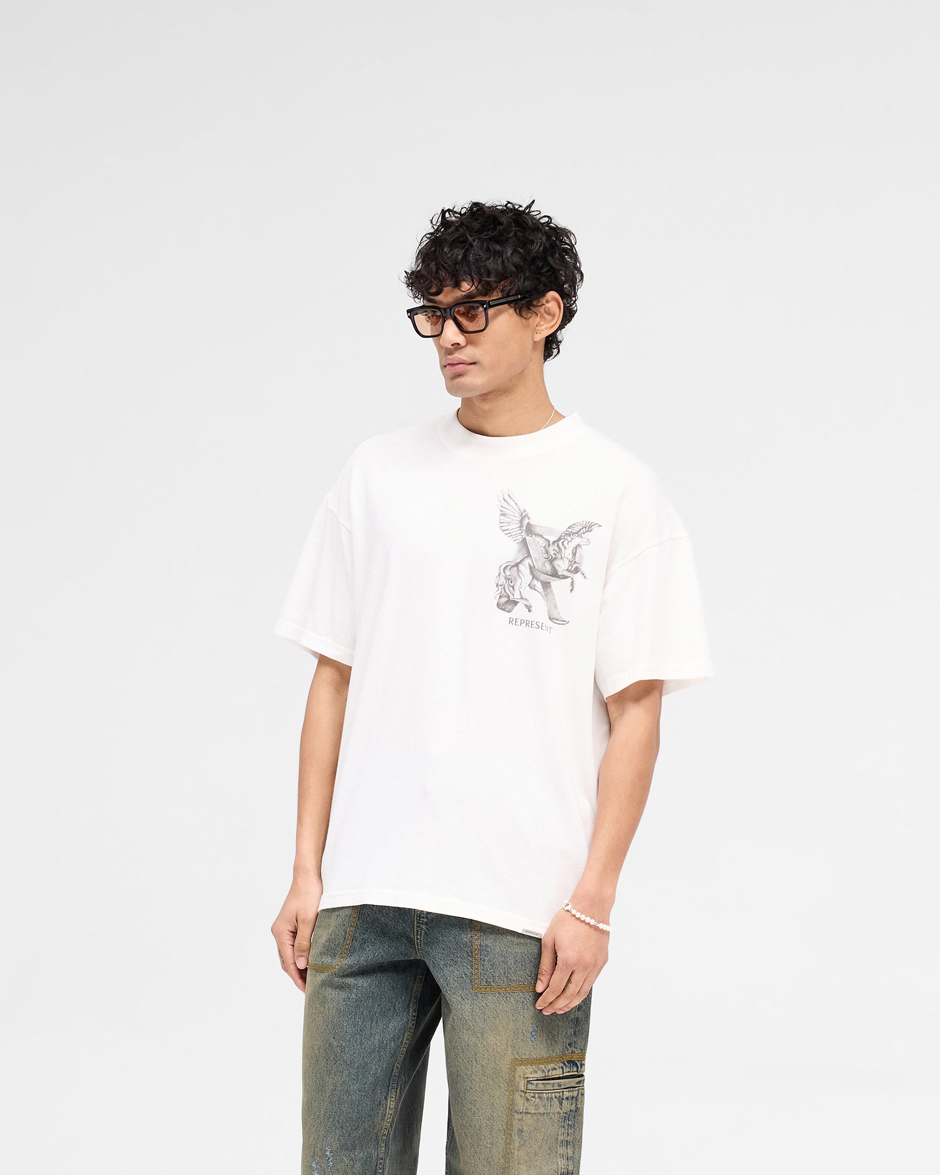 Man wearing a white graphic t-shirt on a plain white background | Elegance In Motion T-Shirt in Flat White | Oversized t-shirt with graphic and metal bar logo | SS25 T-Shirts | Represent Clo