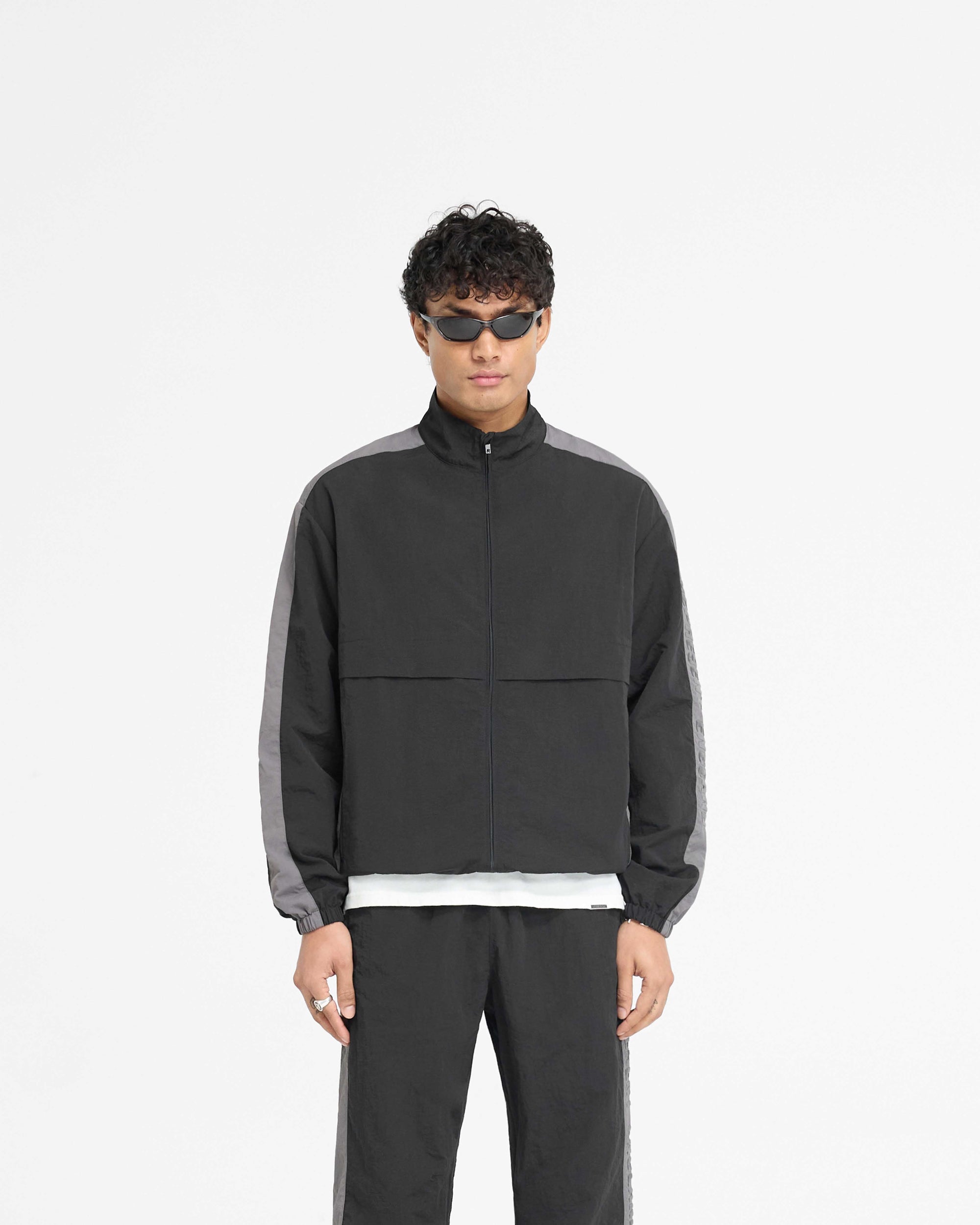 Man wearing a black nylon track jacket on a plain white background | Nylon Track Jacket in Black | Tracksuit jacket with storm flaps and crinkle texture fabric | PRE SS25 Outerwear | Represent Clo