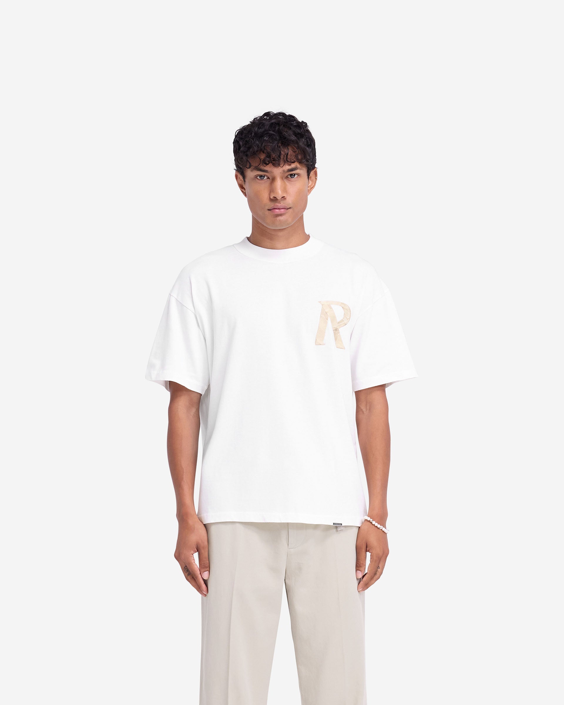 Man wearing a white t-shirt with 3D initial detail on a plain white background | Masking Tape Initial T-Shirt in Flat White | Slightly oversized luxury t-shirt with Represent metal bar | FW24 T-Shirts | Represent Clo