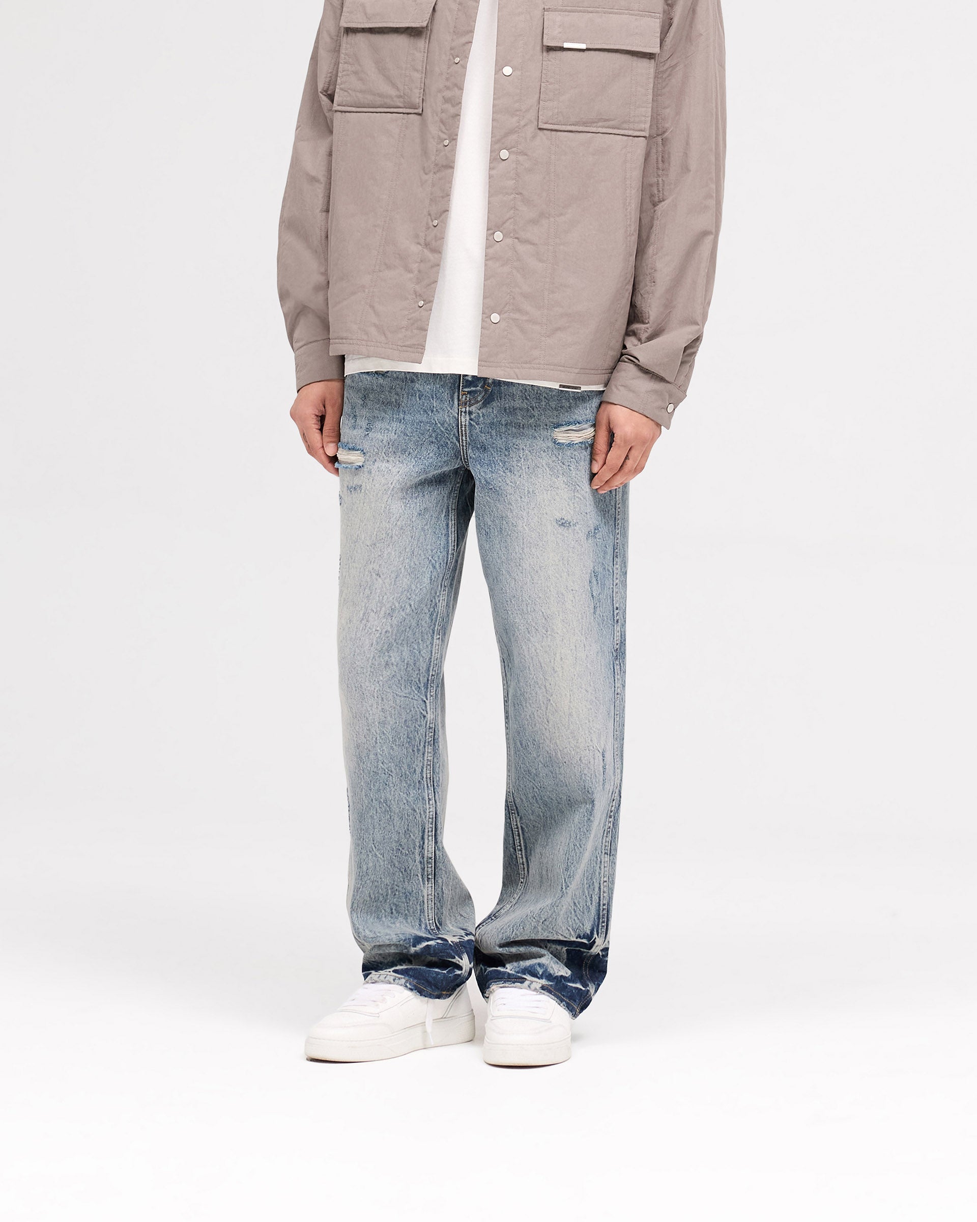 Man wearing distressed baggy blue denim on a plain white background | R3 Distressed Baggy Denim in Blue Cream | Baggy fit jeans with ripped details and initial embroidery | SS25 Denim | Represent Clo