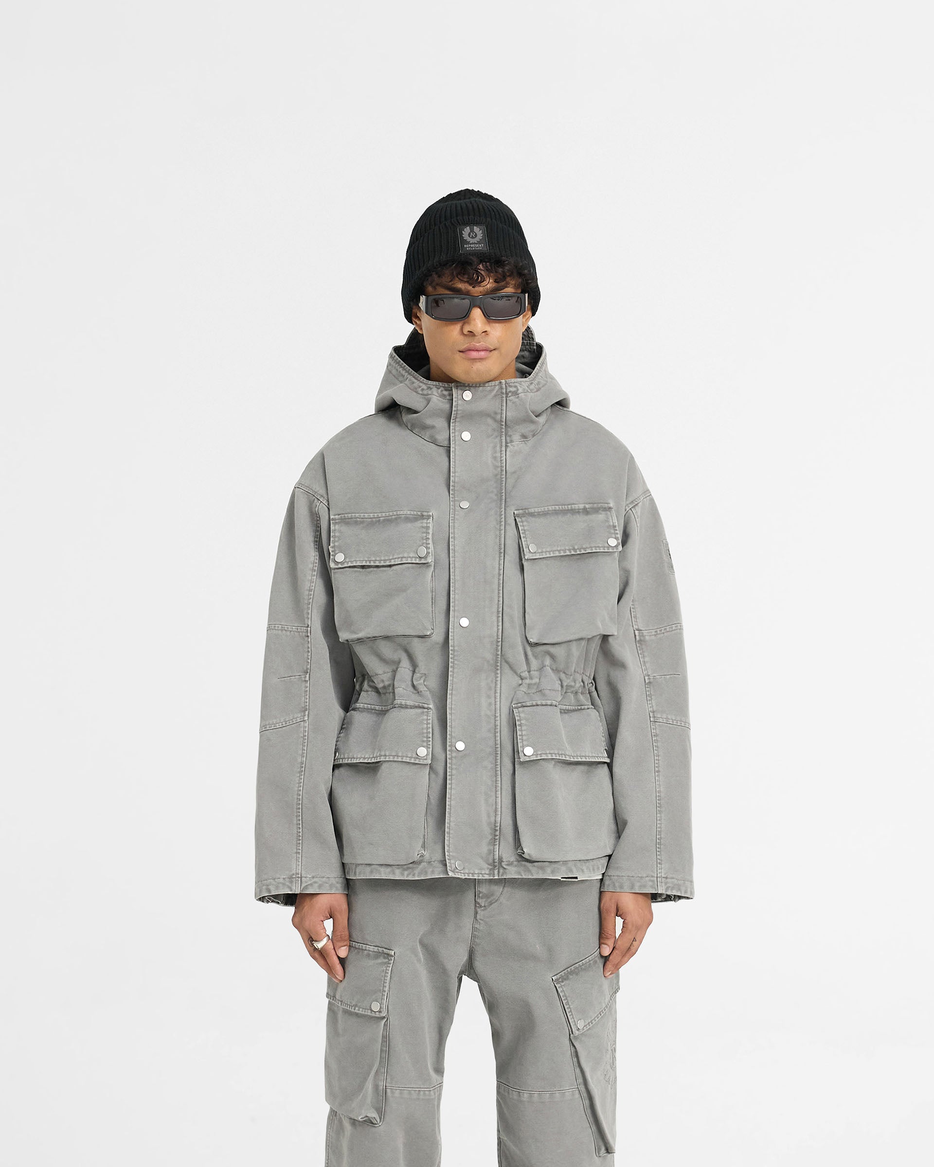 Man wearing a grey field jacket on a plain white background | Represent X Belstaff Enduro Jacket Shadow | Oversized cotton field jacket with adjustable hood and pockets | Exclusives Outerwear | Represent Clo
