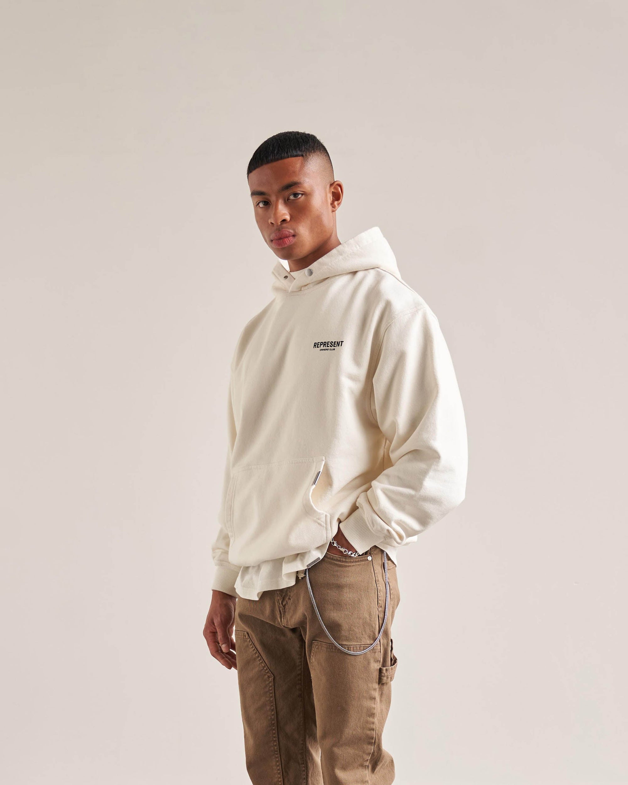 Man wearing an oversized white hoodie on a plain white background | Represent Owners Club Hoodie in Flat White | Oversized fit hoodie with Cobrax popper detail | Owners Club Hoodies | Represent Clo
