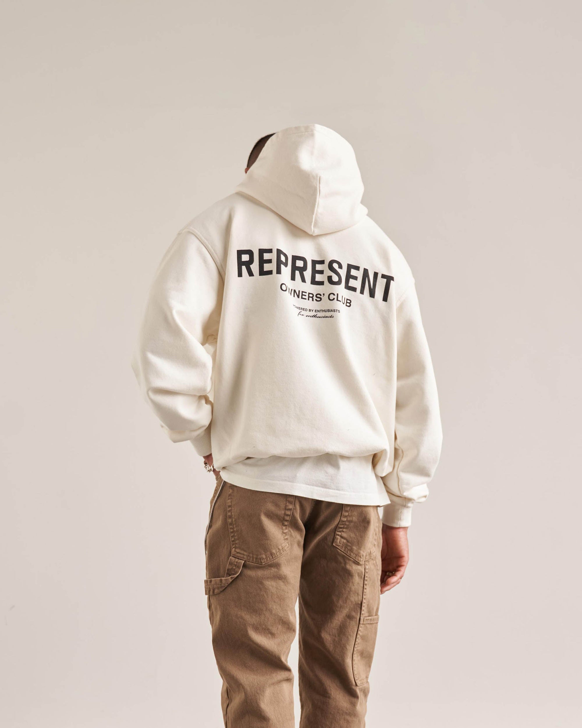 Man wearing an oversized white hoodie on a plain white background | Represent Owners Club Hoodie in Flat White | Heavyweight jersey hoodie with Owners Club print | Owners Club Hoodies | Represent Clo