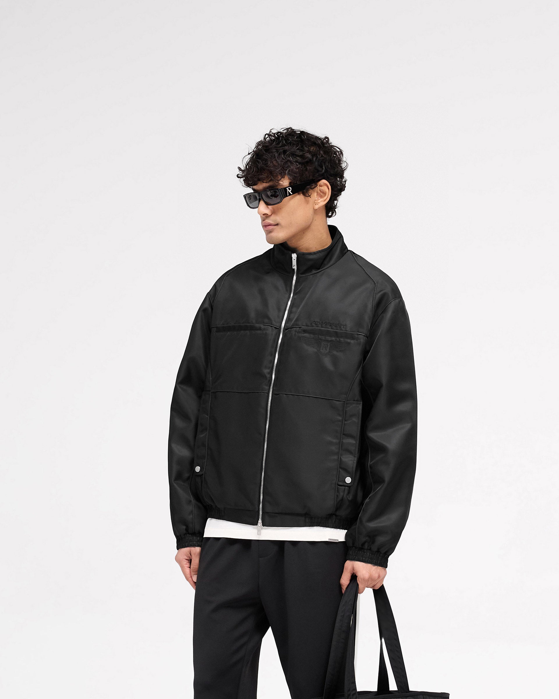 Man wearing an oversized black jacket on a plain white background | Overdrive Racing Jacket in Black | Racing jacket with funnel neck and double ended zipper | SS25 Outerwear | Represent Clo