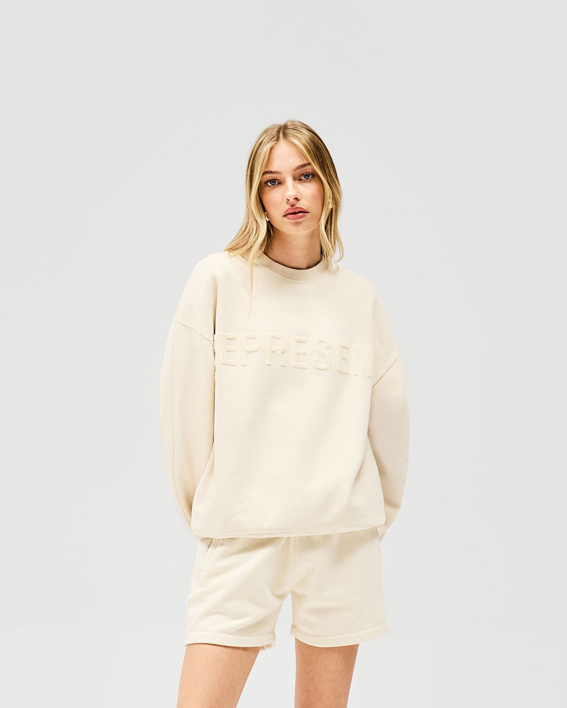Woman wearing a relaxed oat milk sweater on a plain white background | Owners Club Sweater in Oat Milk | Sweater with ribbed hem and Represent branding | Womens Owners Club Sweaters | Represent Clo