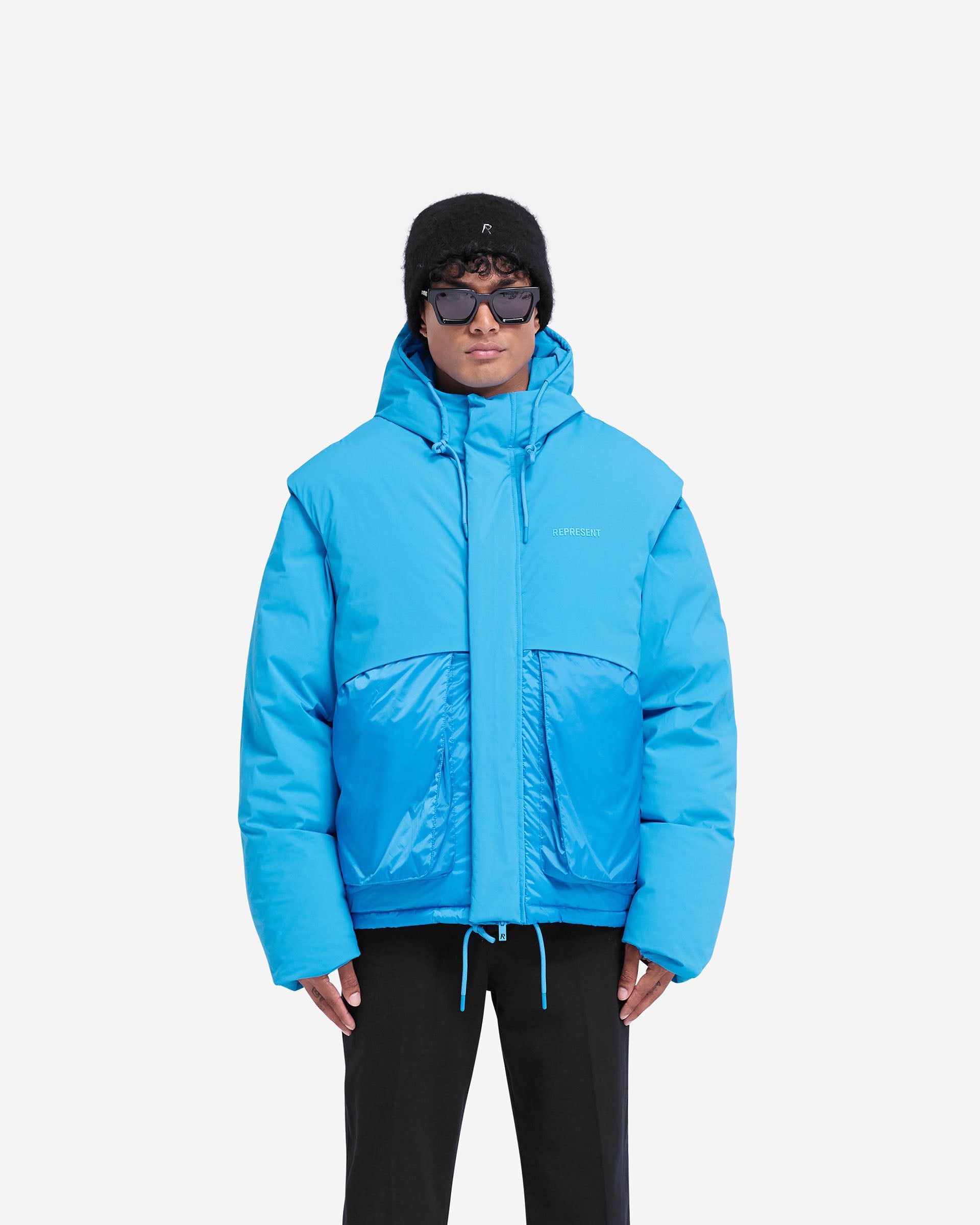 Man wearing a blue puffer jacket on a plain white background | Layered Hooded Puffer in Electric Blue | Duck down filled jacket with adjustable hood and storm flaps | FW24 Outerwear | Represent Clo