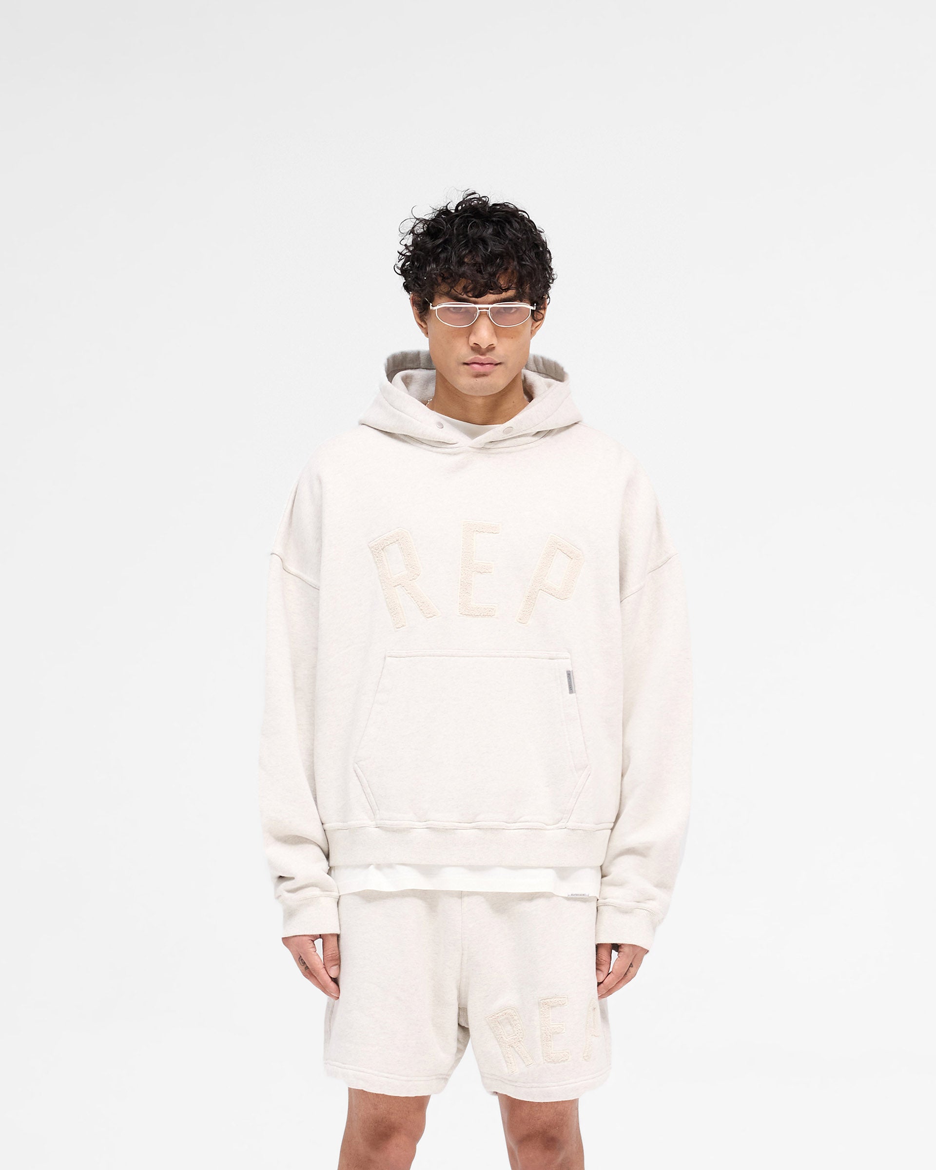 Man wearing a cream hoodie on a plain white background | Rep Applique Hoodie in Cream Marl | Boxy fit hoodie with textured Rep applique | SS25 Hoodies | Represent Clo