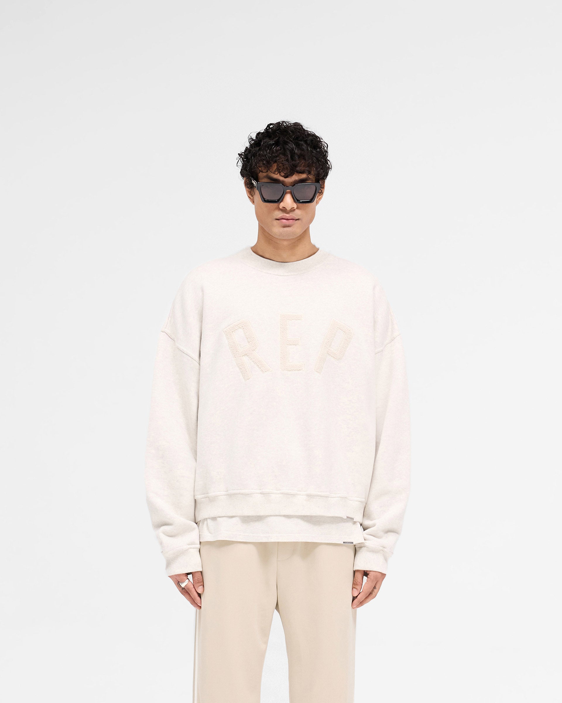 Man wearing a cream sweater on a plain white background | Rep Applique Sweater in Cream Marl | Boxy fit sweater with textured rep applique | SS25 Sweaters | Represent Clo