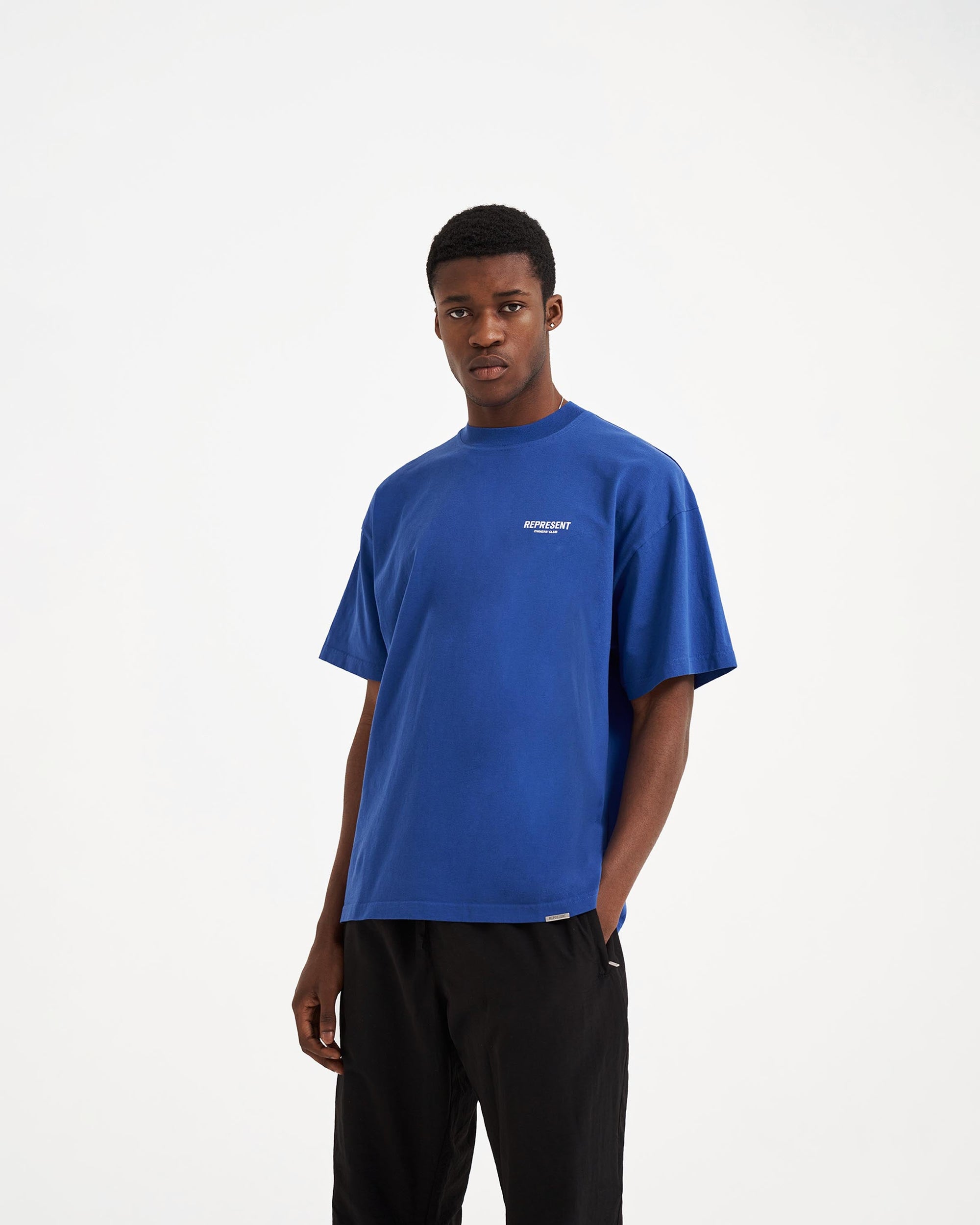 Man wearing a cobalt blue t-shirt on a plain white background | Represent Owners Club T-Shirt in Cobalt | Oversized fit cotton t-shirt with ribbed crewneck and graphic branding | Owners Club T-Shirts | Represent Clo