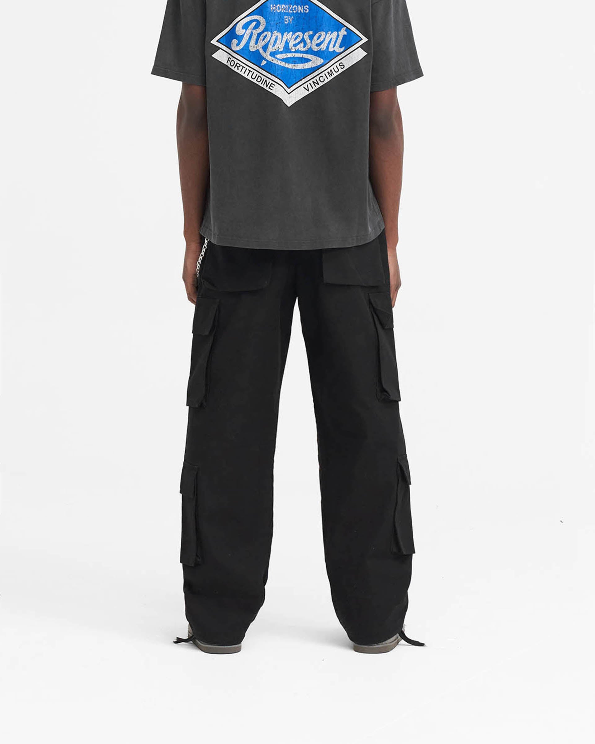 Man wearing black baggy cargo pants on a plain white background | Baggy Cargo Pant in Black | Cargo pants with 3D oversized pockets and antique silver hardware | SS24 Pants | Represent Clo
