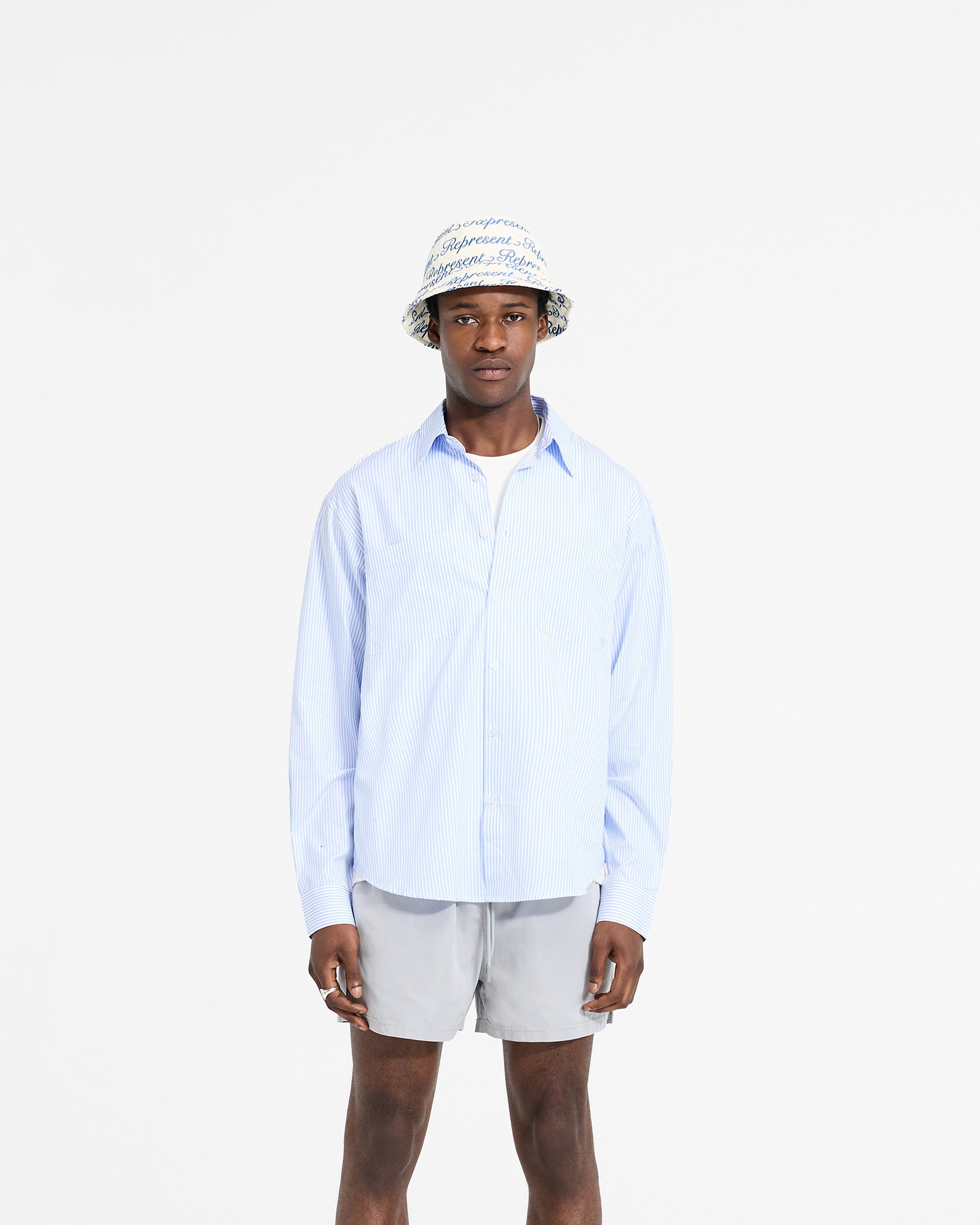 Man wearing a blue pinstripe long sleeve shirt on a plain white background | Long Sleeve Pinstripe Shirt in Baby Blue | Slightly oversized fit shirt with curved hem and concealed placket | SC24 Shirts | Represent Clo