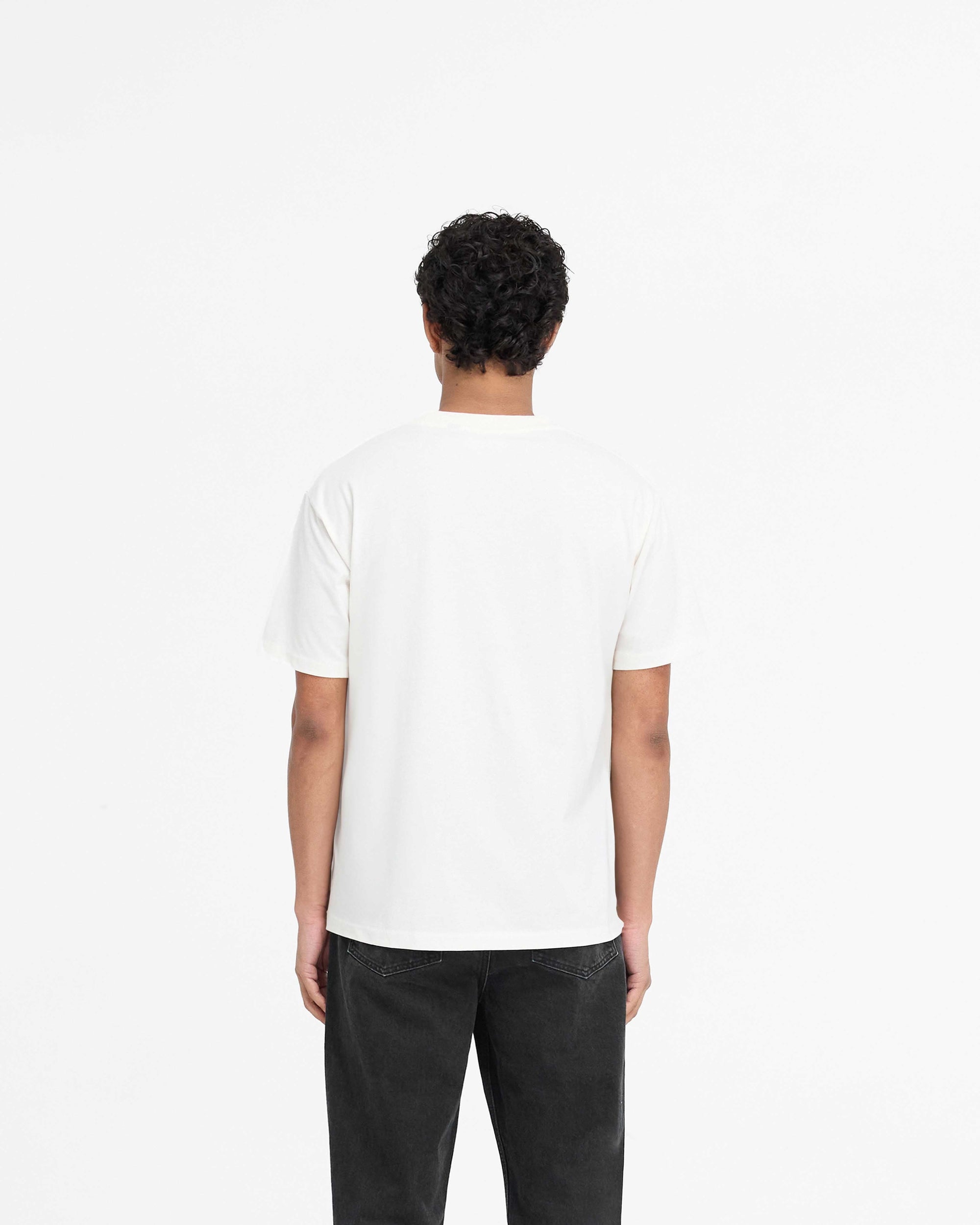 Man wearing a slightly oversized white t-shirt on a plain white background | Script Logo T-Shirt in Flat White | Super soft t-shirt with high build embroidered script and metal bar logo | PRE SS25 T-Shirts | Represent Clo