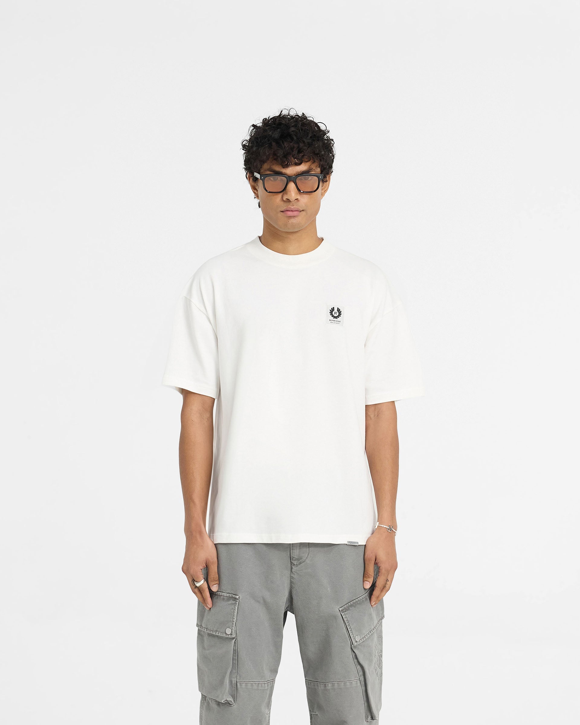 Man wearing a white t-shirt on a plain white background | Represent X Belstaff Patch T-Shirt in Flat White | Oversized t-shirt with woven patch branding | Exclusives T-Shirts | Represent Clo