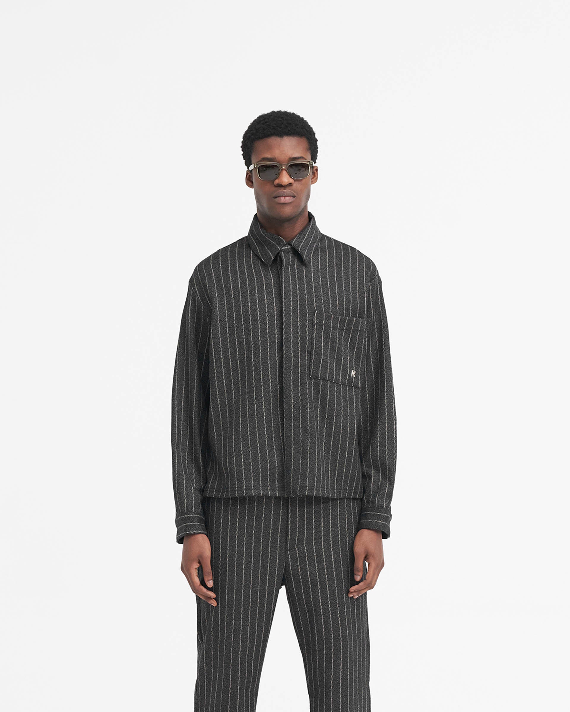 Man wearing a black pinstripe cropped dress shirt on a plain white background | Cropped Dress Shirt in Black Pinstripe | Dress shirt with stepped hem and Cobrax poppers | SS24 Shirts | Represent Clo