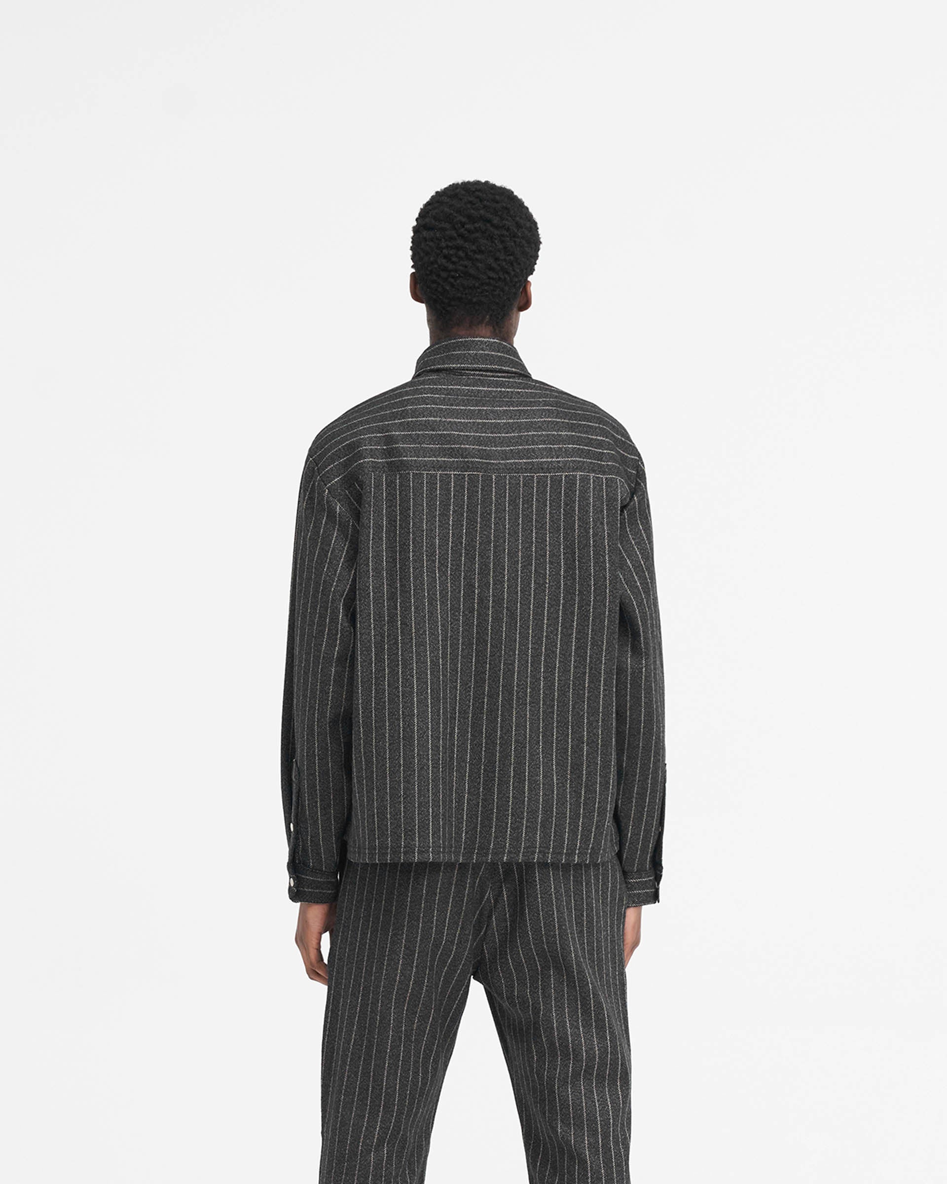 Man wearing a black pinstripe cropped dress shirt on a plain white background | Cropped Dress Shirt in Black Pinstripe | Dress shirt with stepped hem and antique silver logo | SS24 Shirts | Represent Clo
