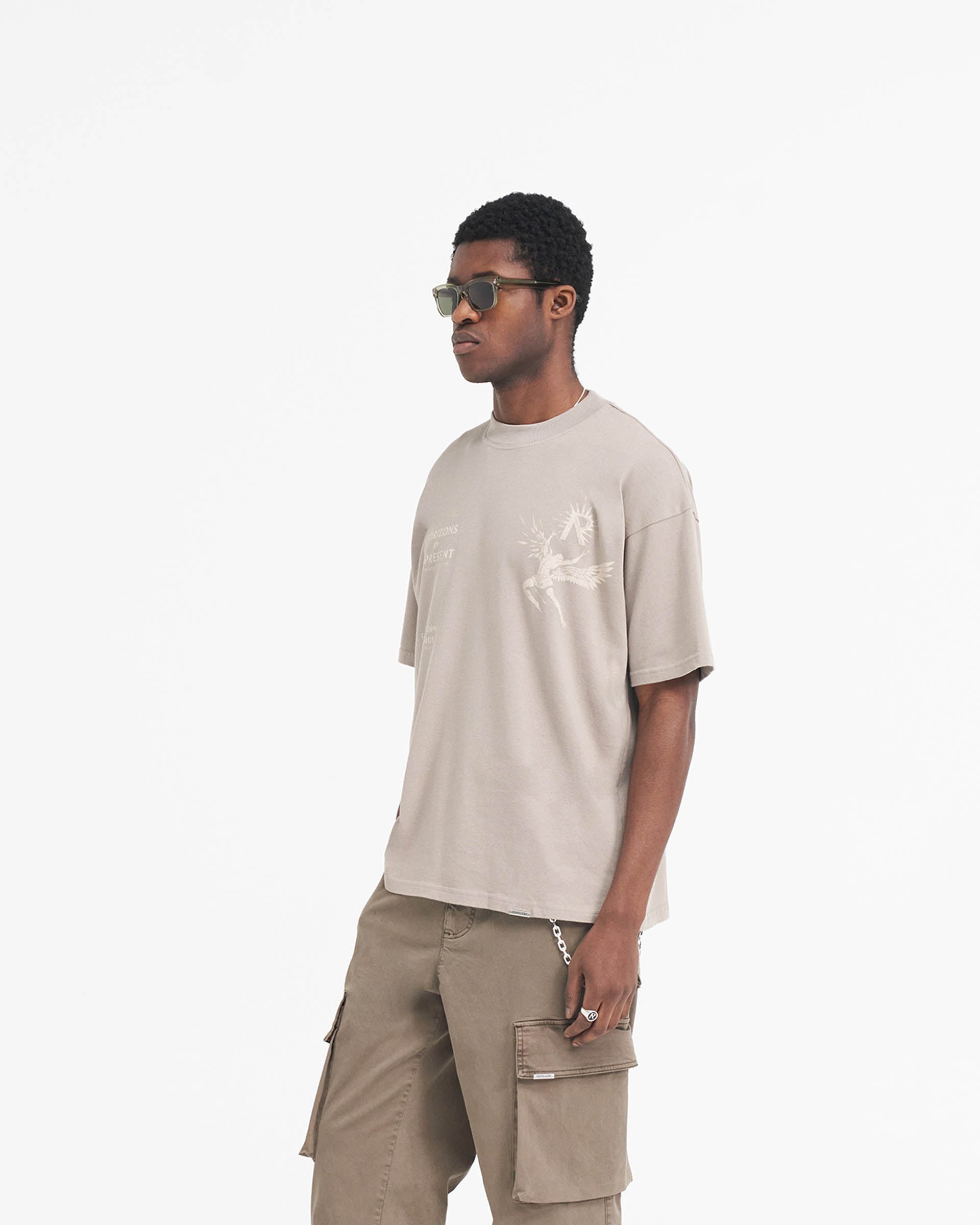 Man wearing an oversized taupe t-shirt with Icarus graphic on a plain white background | Icarus T-Shirt in Taupe | Short sleeves round neck t-shirt with Greek mythology inspired design | SS24 T-Shirts | Represent Clo