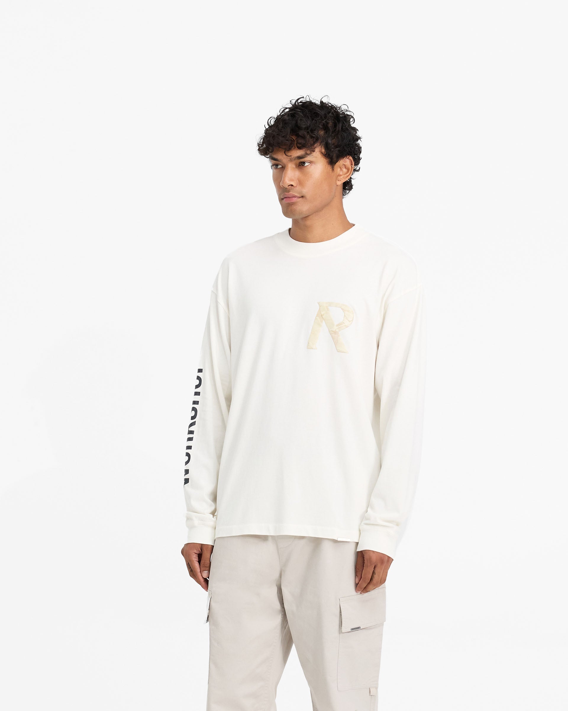 Man wearing a white oversized long sleeve t-shirt on a plain white background | Masking Tape Initial Long Sleeve T-Shirt in Flat White | Long sleeve t-shirt with 3D masking tape effect | FW24 T-Shirts | Represent Clo
