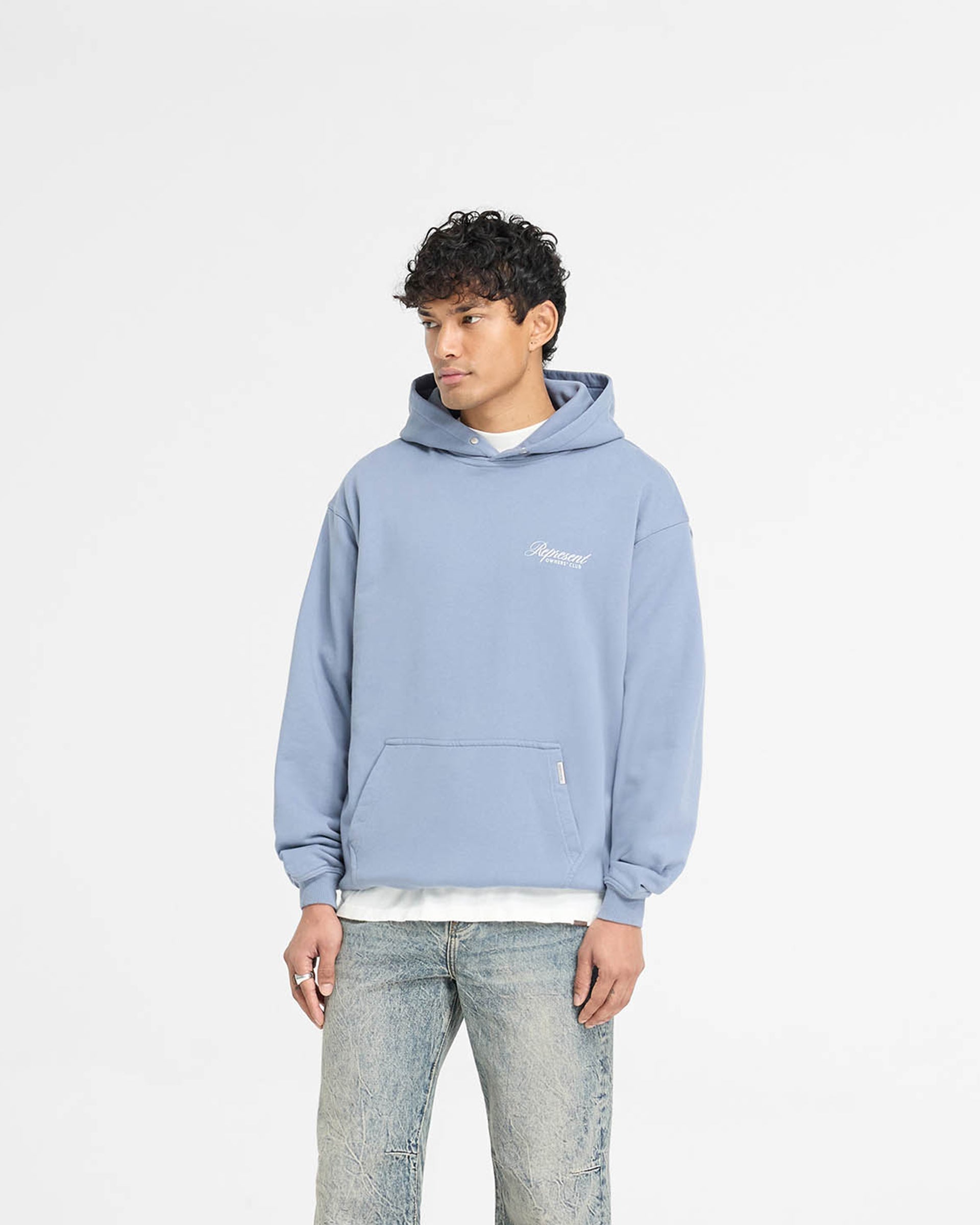 Man wearing a dusty blue hoodie on a plain white background | Represent Owners Club Script Hoodie in Dusty Blue | Heavy weight oversized hoodie with screen print graphic | Owners Club Hoodies | Represent Clo
