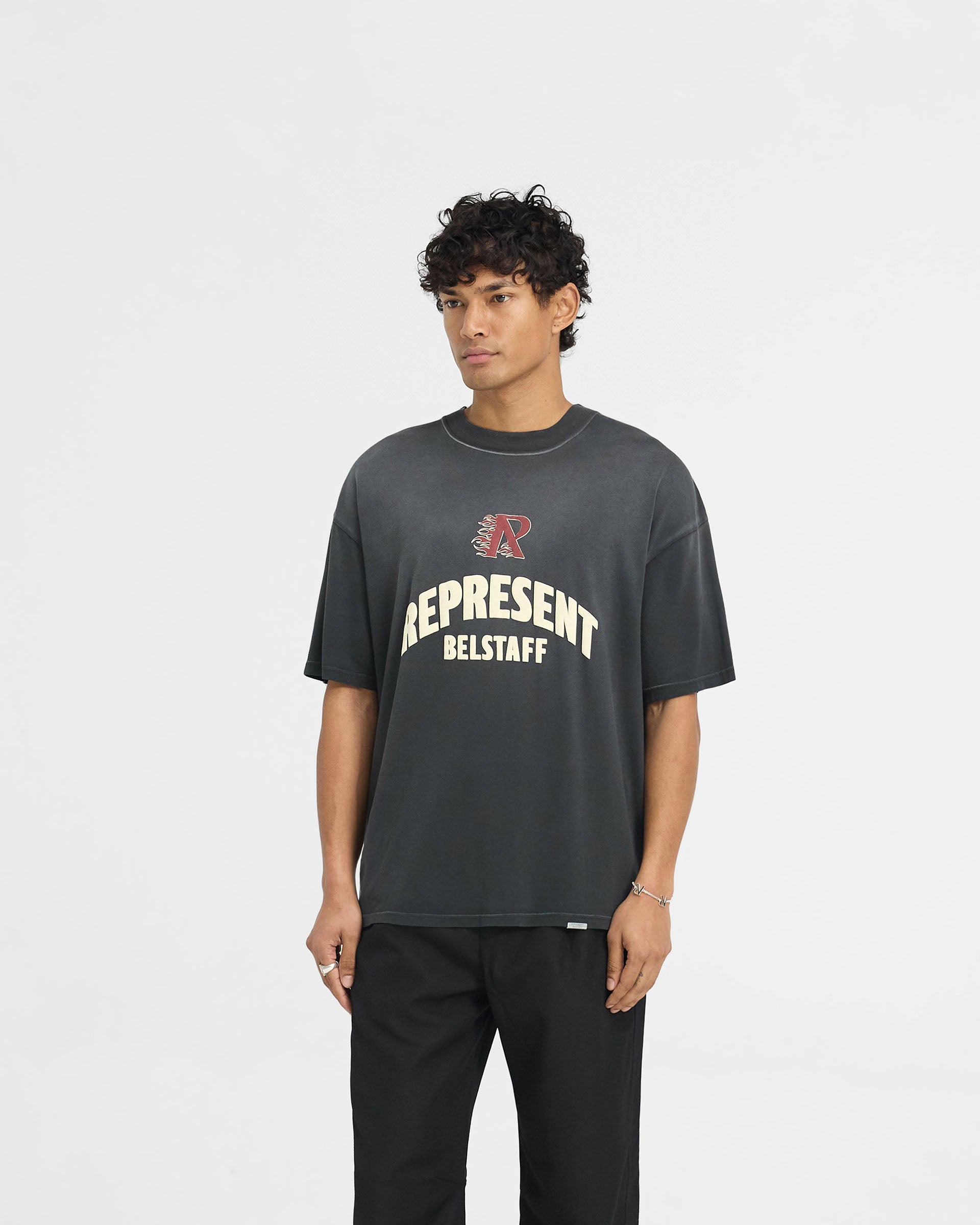 Man wearing an oversized black t-shirt on a plain white background | Represent X Belstaff Flame Phoenix T-Shirt in Shadow | Medium weight t-shirt with Phoenix and Initial logos | Exclusives T-Shirts | Represent Clo