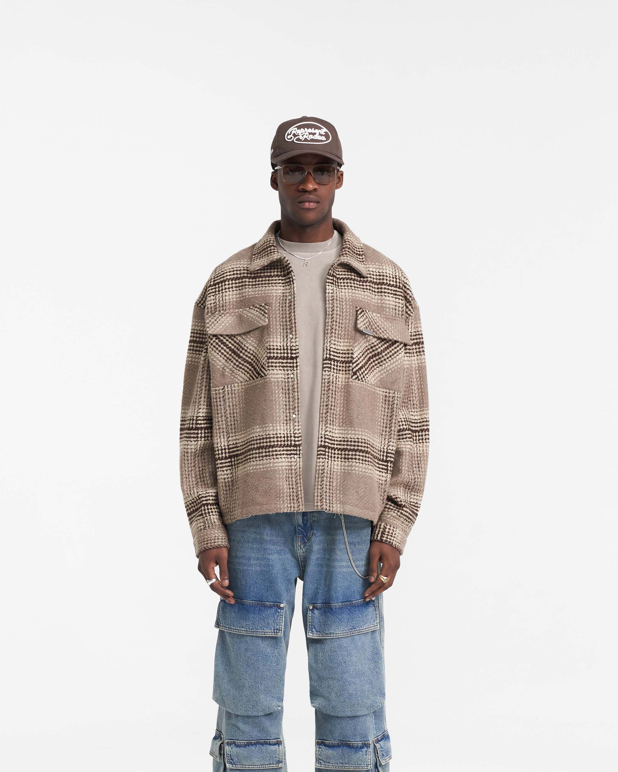 Man wearing a brown check overshirt on a plain white background | Represent Flannel Shirt in Brown Check | Overshirt with high build embroidery and chest patch pockets | FW24 Shirts | Represent Clo