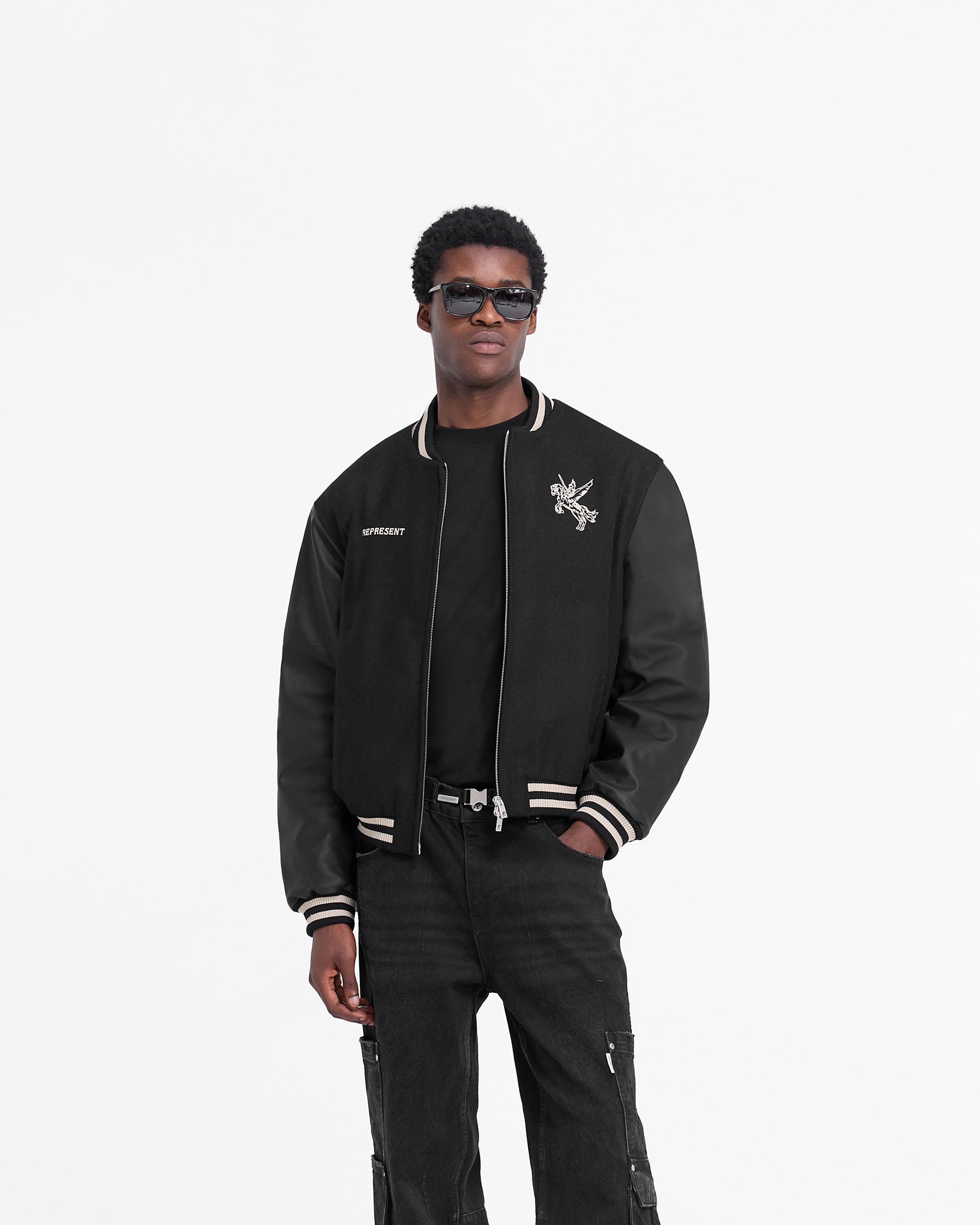 Man wearing a black varsity jacket on a plain white background | Mascot Wool Varsity Jacket in Jet Black | Oversized varsity jacket with embroidered mascot and PU sleeves | FW24 Outerwear | Represent Clo